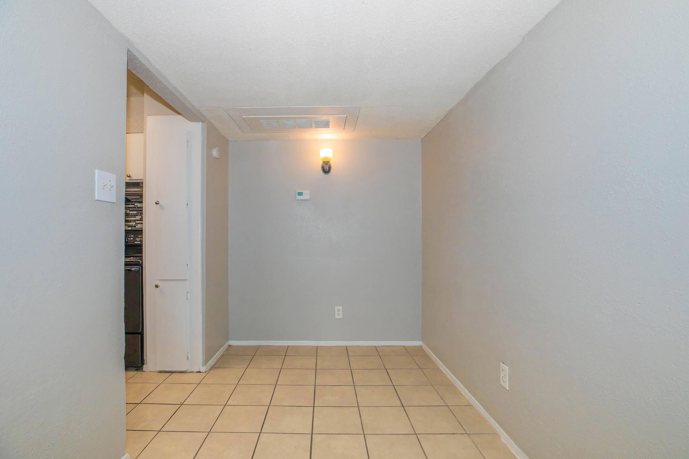 Empty room with light gray walls and a tiled floor. A small wall-mounted light fixture is visible, along with a thermostat on the wall. A doorway leads to another space, and there are no furnishings in the room. The overall appearance is clean and minimalistic.