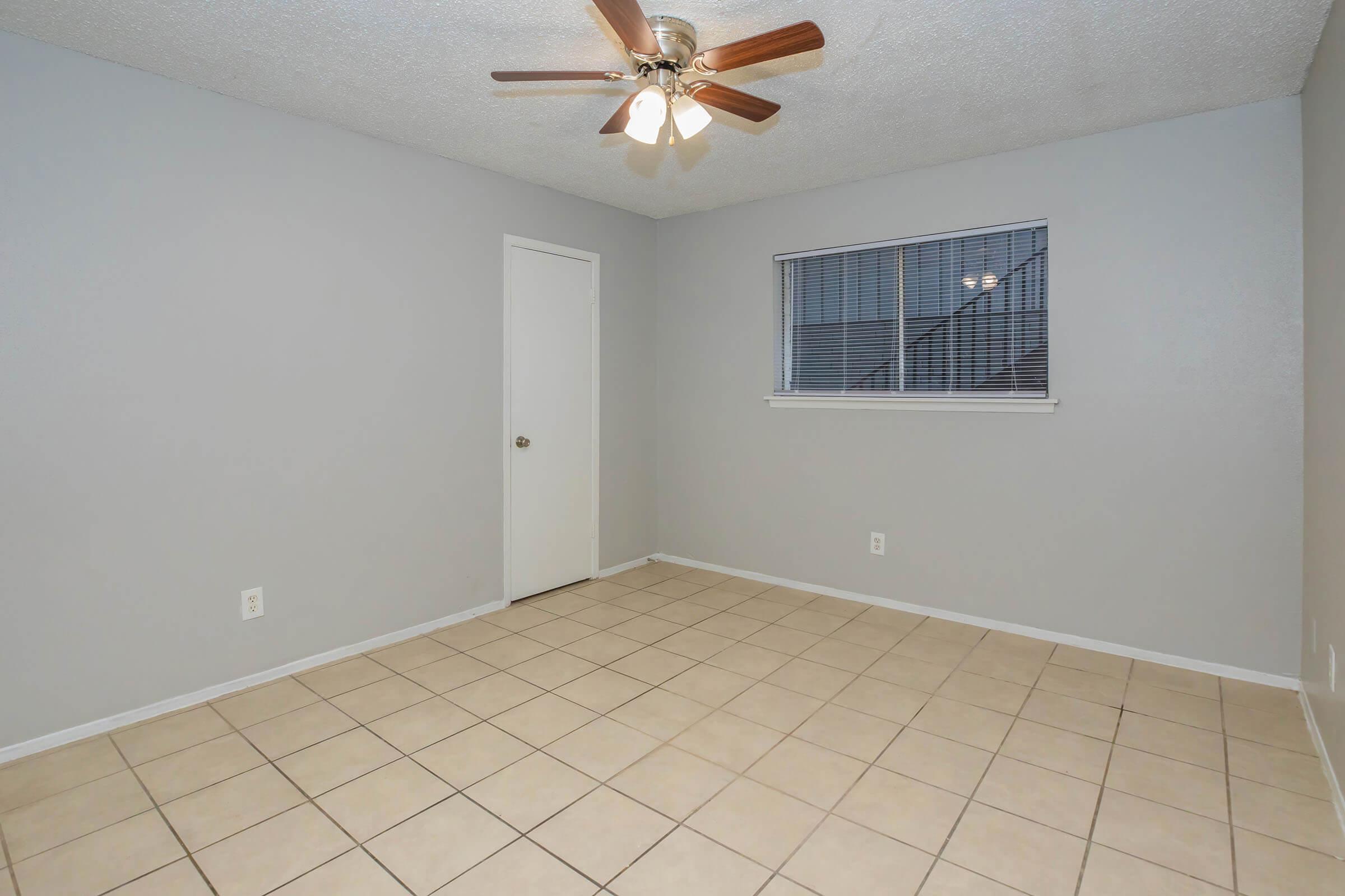 Empty room featuring light gray walls and tile flooring. A ceiling fan with wooden blades hangs from the ceiling, providing light. There is a closed door to the left and a window with blinds on the right, allowing natural light. The space is simple and uncluttered, suitable for various purposes.