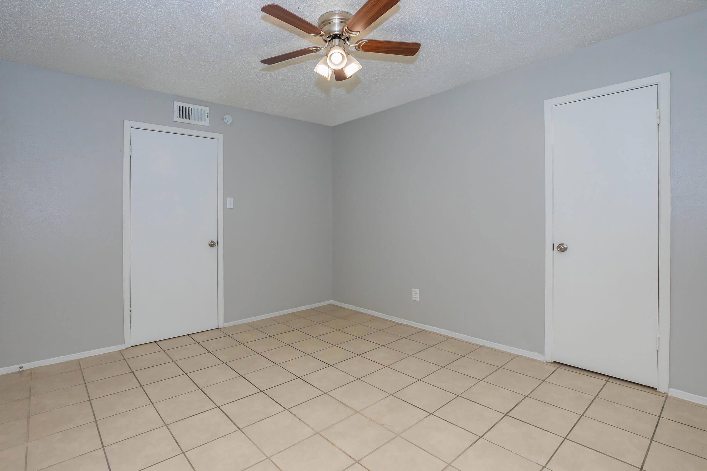 A small, empty room with light gray walls and a ceiling fan. The floor is tiled with square beige tiles. There are two white doors on opposite walls, and the room has no furniture, creating an open and spacious feel.