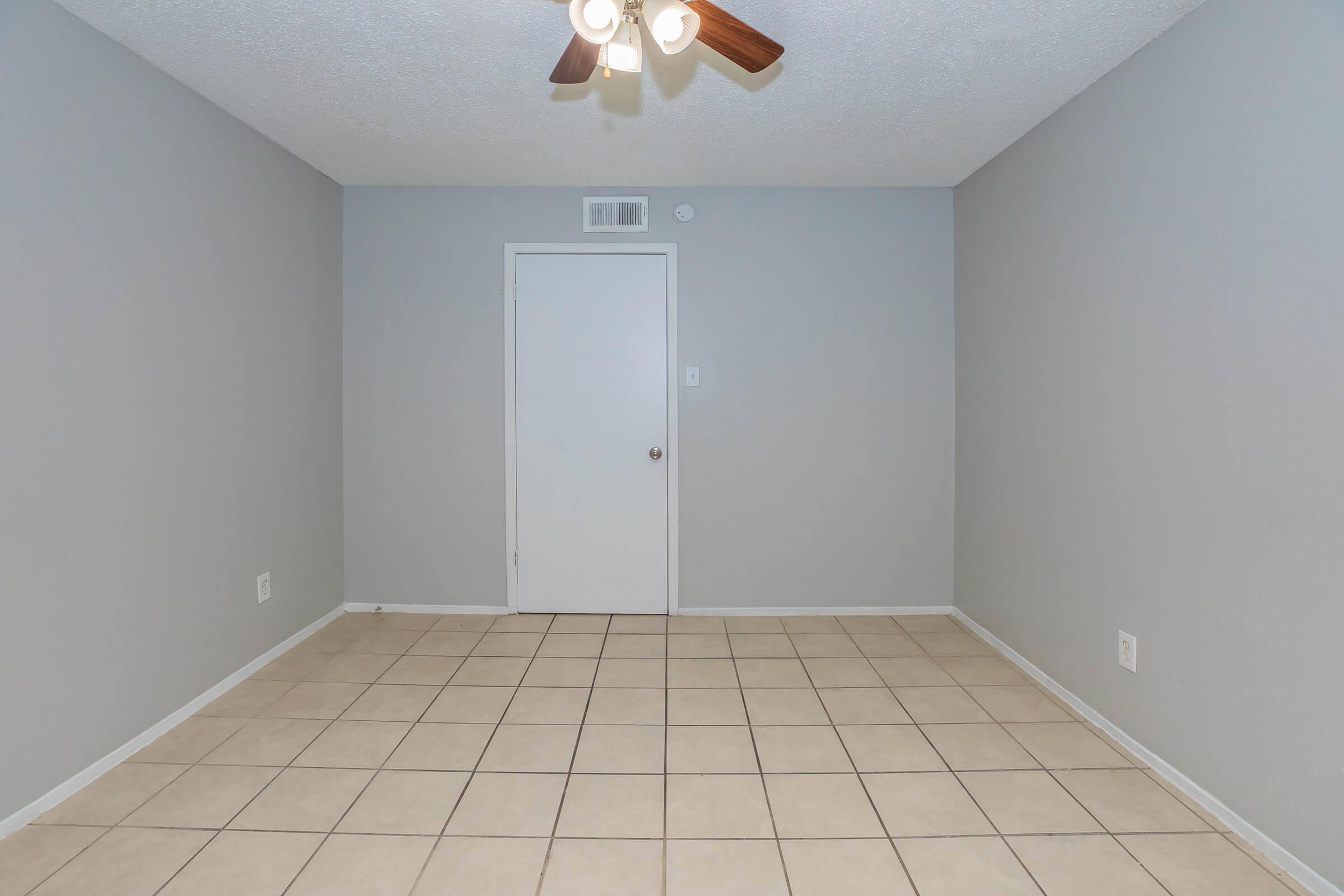 Empty room with gray walls and a ceiling fan. The floor is tiled, and there is a closed door on one wall. Natural light illuminates the space, creating a clean and neutral atmosphere. Suitable for various uses such as a living area or office.