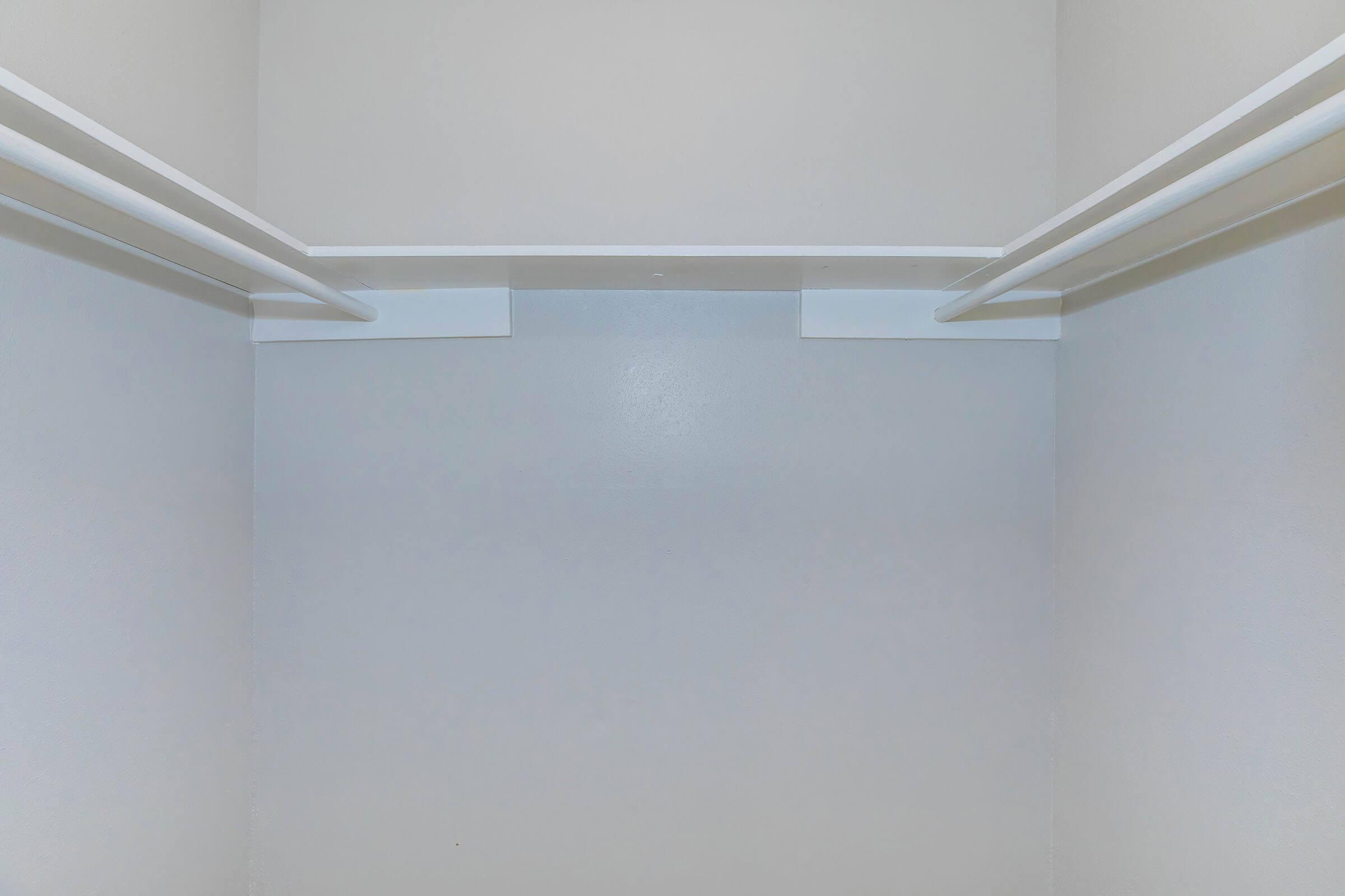 Empty closet interior with plain white walls and two horizontal clothing rods positioned near the top. The floor is also bare, contributing to the minimalistic and spacious appearance. Light reflects softly off the surfaces, emphasizing the simplicity of the design.