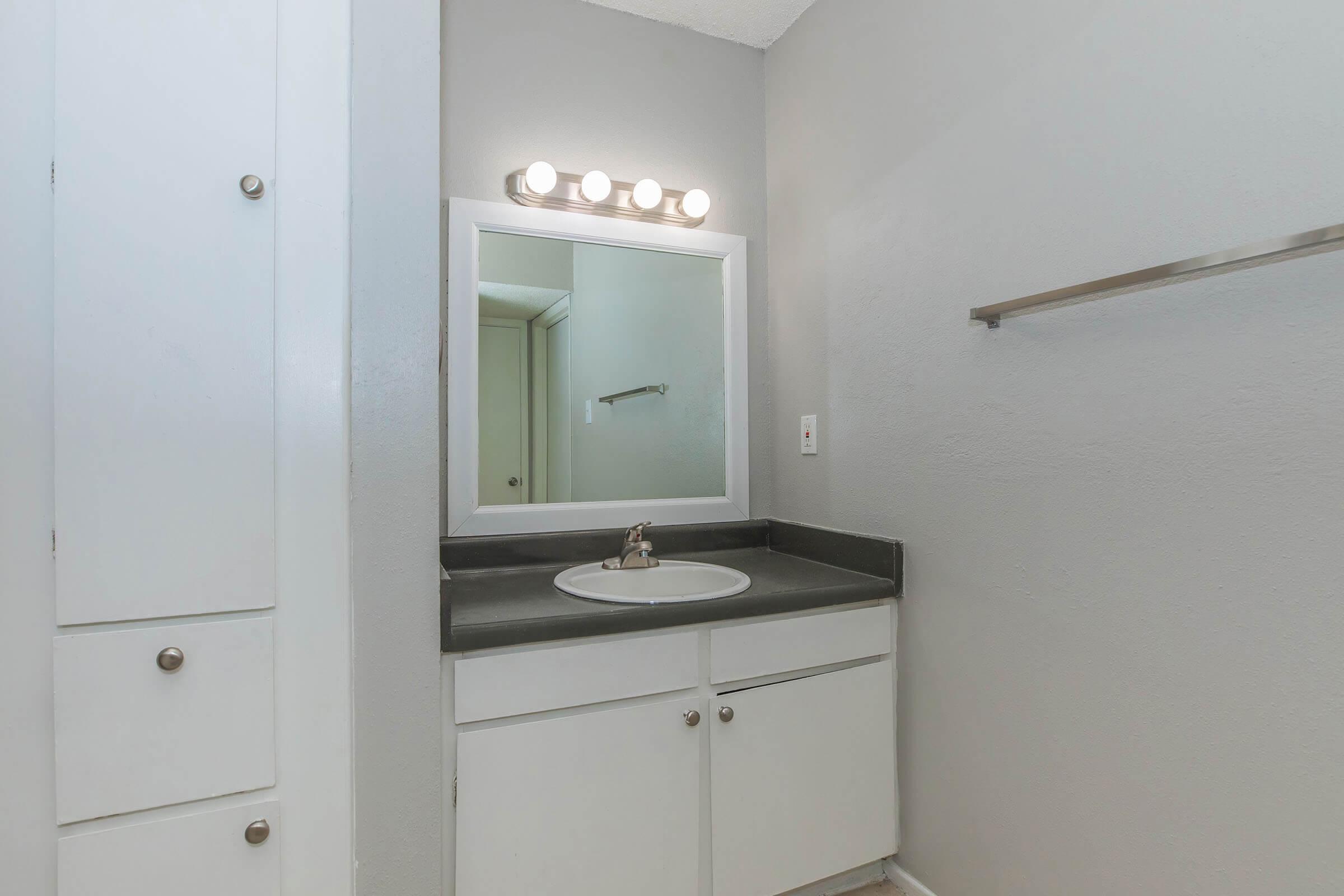 A simple bathroom featuring a small sink with a faucet on a dark countertop. A mirror is above the sink, and there are light fixtures above it. The walls are painted gray, and there's a towel bar on the right. A cabinet with white doors is visible on the left side of the image.