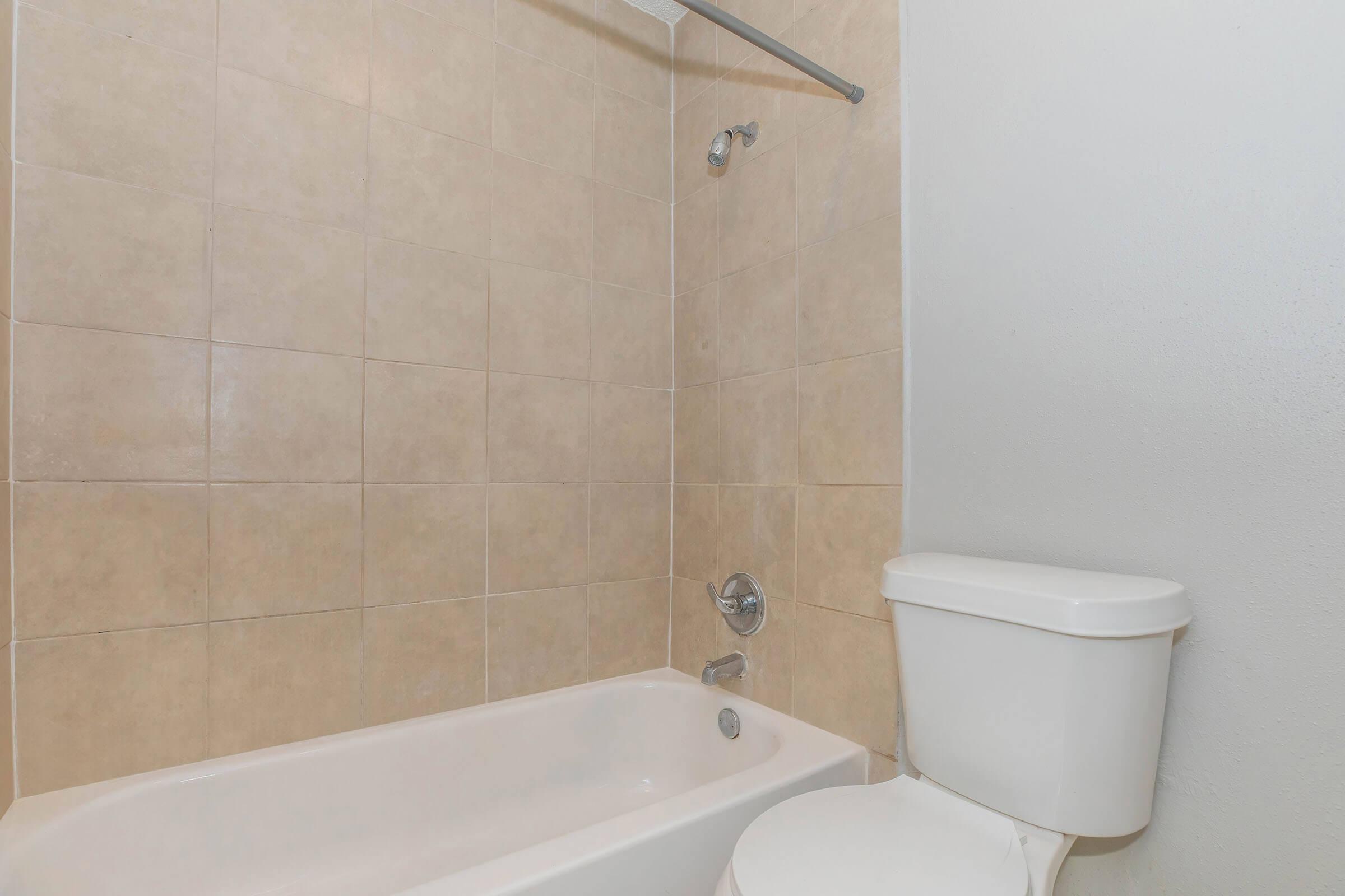 A clean bathroom featuring a beige tiled shower and bathtub combination, with a chrome showerhead and controls. Next to the bathtub, there is a white toilet. The walls are painted a light color, providing a spacious and simple aesthetic.
