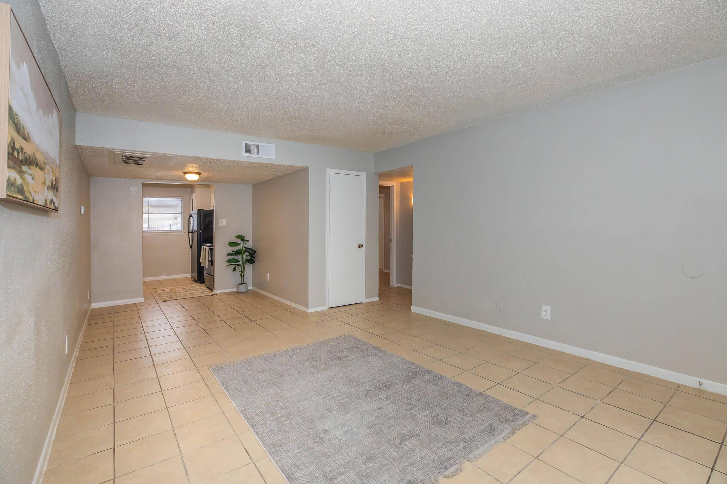 A spacious, empty living area with beige tile flooring and a neutral gray wall color. A small area rug is placed on the floor. To the left, a doorway leads to a kitchen, partially visible with dark cabinets and appliances. The room is well-lit with natural light coming from a window.