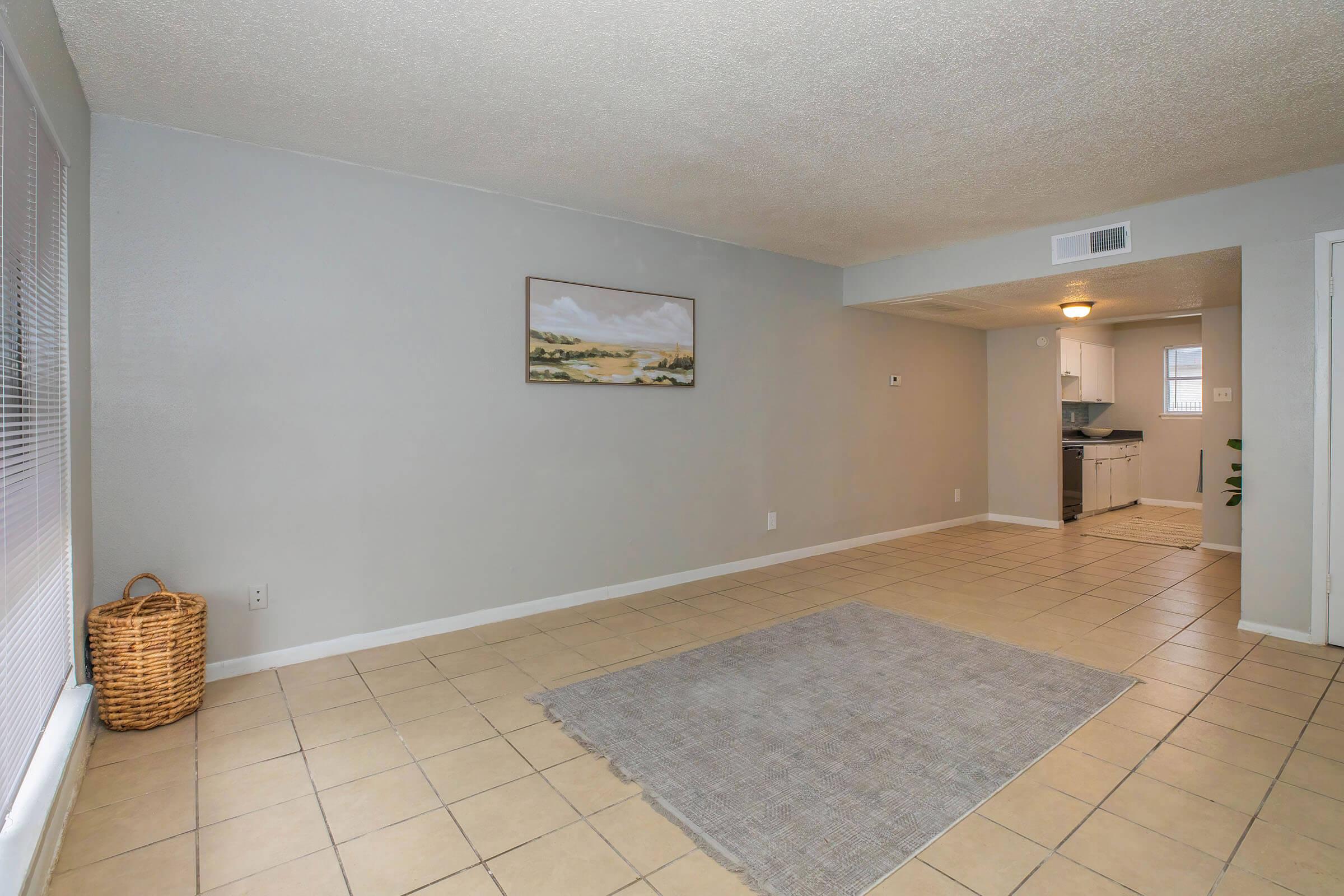 A spacious, empty living room featuring light gray walls and beige tile flooring. A large area rug is in the center, and a woven basket sits in the corner. A painting of a landscape is hung on the wall. The room leads into a kitchen area, partially visible in the background.