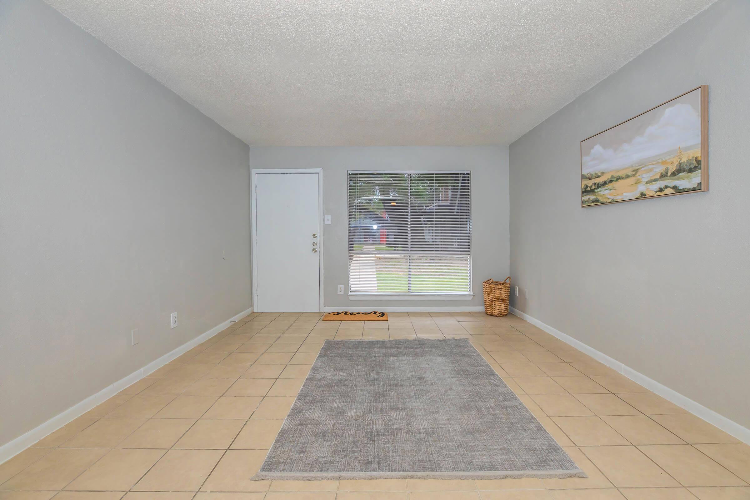 A spacious, light gray living room with tiled flooring. There is a rectangular gray area rug in the center, a large window providing natural light, and a door leading outside. A wall features a framed landscape painting, and a woven basket is visible in the corner.
