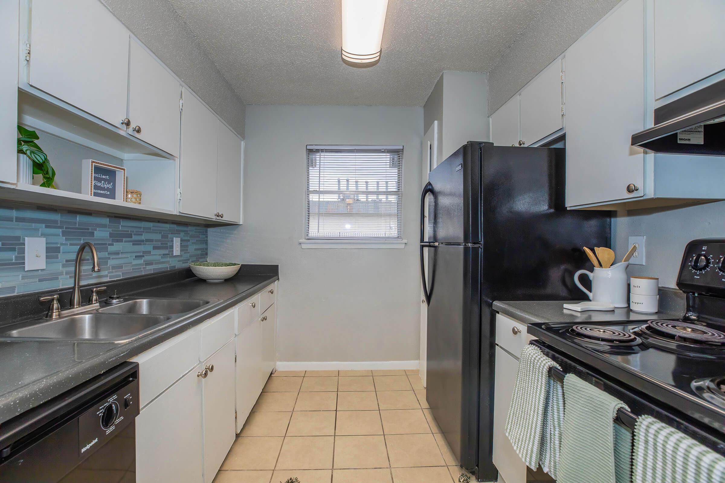 A modern kitchen featuring light-colored cabinets, a black refrigerator, and a black stove. The countertop is dark, and there's a tiled backsplash. Natural light comes through a window. The floor is tiled, and kitchen accessories are visible, including a bowl and utensils.