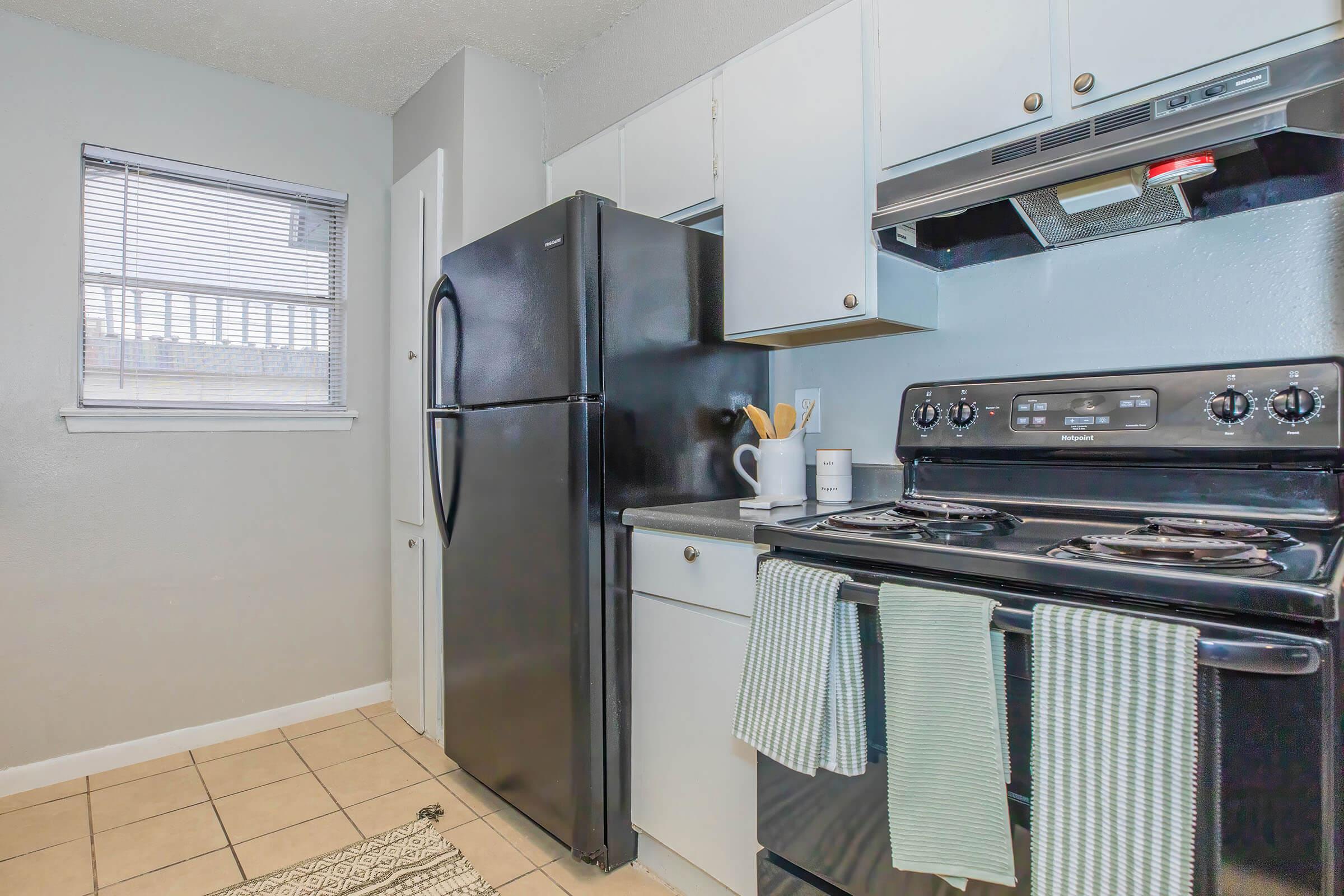 A modern kitchen featuring a black refrigerator, a gray stove with an oven, white cabinets, and a window with blinds. A light-colored rug adds warmth to the tiled floor. Kitchen utensils and accessories are neatly arranged on the countertop, enhancing the inviting atmosphere.