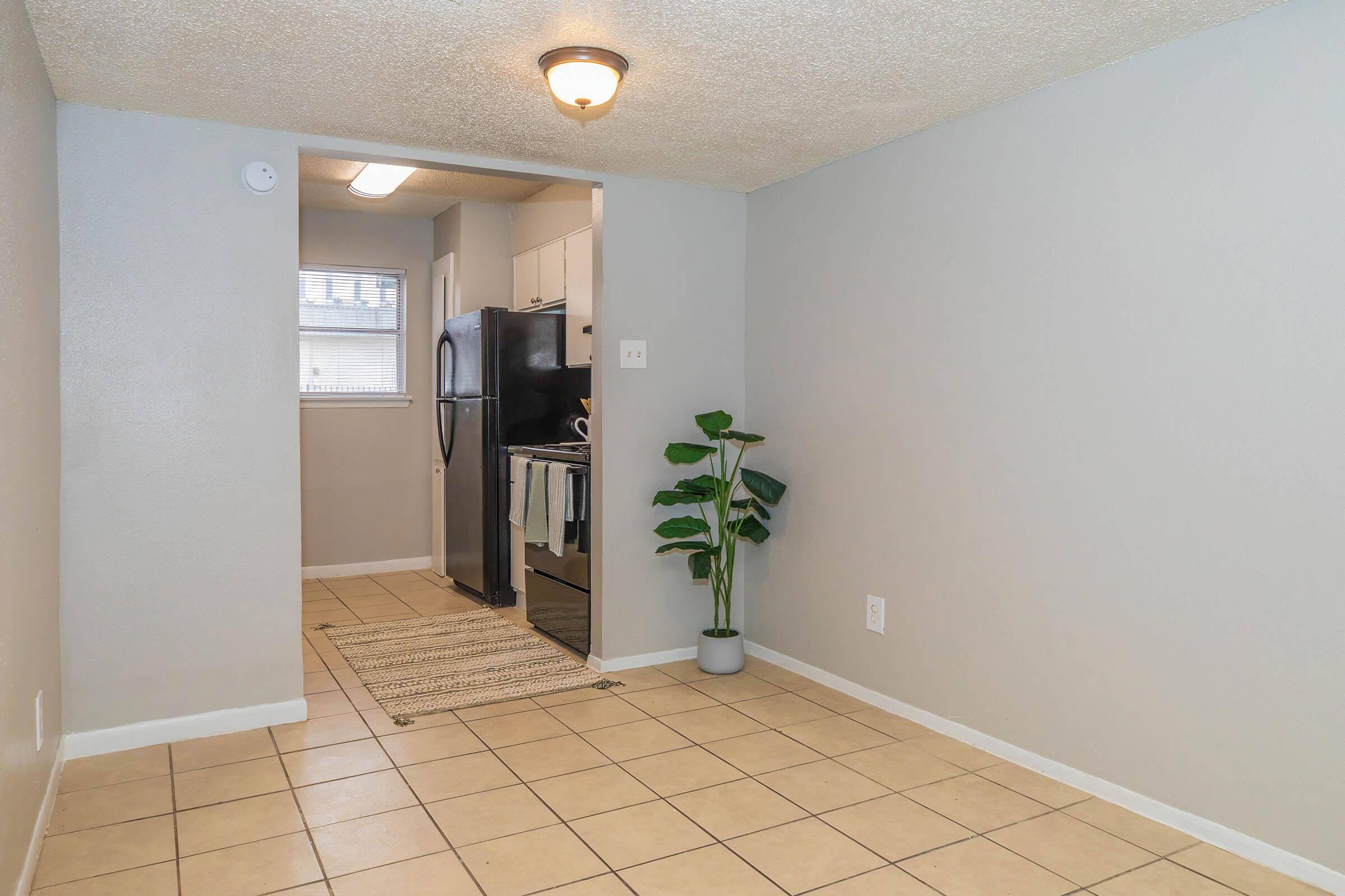 A small, vacant apartment living area featuring light gray walls and tiled flooring. In the background, a kitchen area is visible with a black refrigerator and white cabinets. A green potted plant stands on the floor, and a beige area rug is laid out on the ground. Natural light enters through a window.