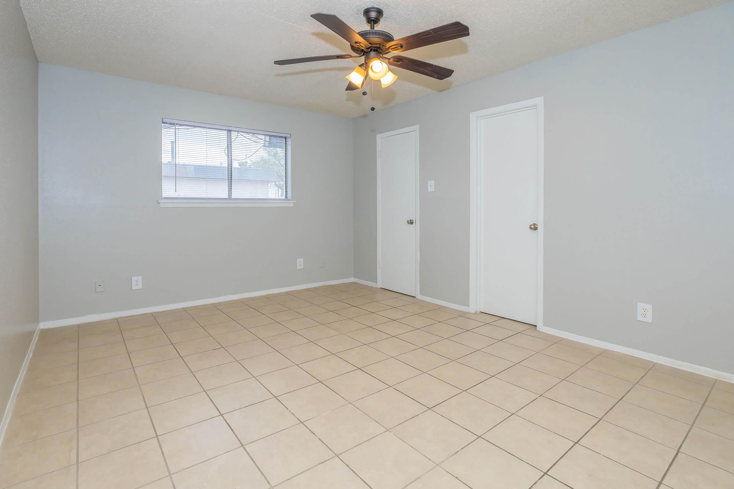 A spacious, empty room with light gray walls and tile flooring. It features a ceiling fan with lights, one window allowing natural light, and two closed doors leading to adjacent areas. The room has a clean and minimalistic look, ideal for various uses.