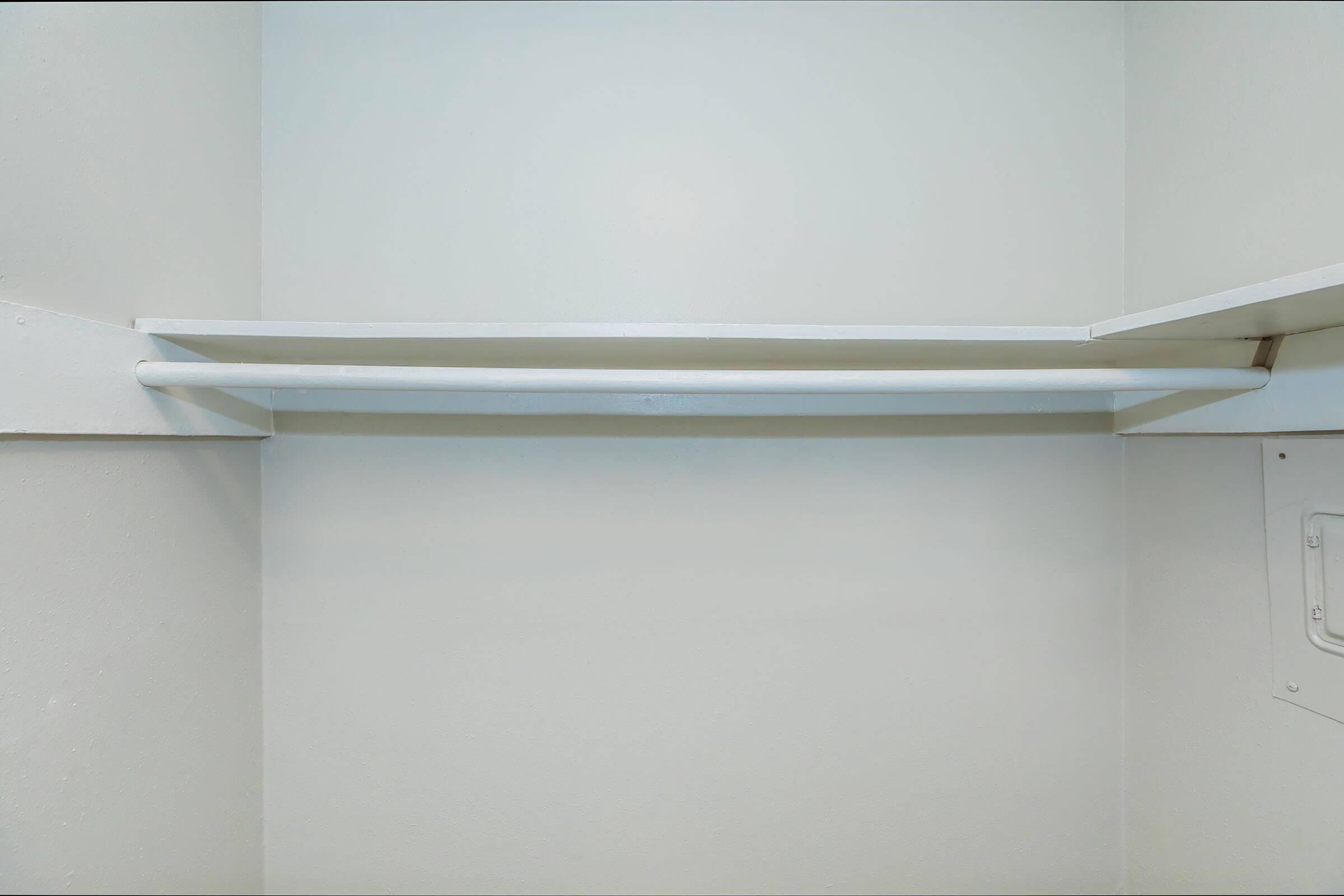Empty closet with smooth, white walls and a single shelf. The shelf is mounted on one side and runs horizontally across the top, with no items stored on it. The overall appearance is minimalistic and tidy.
