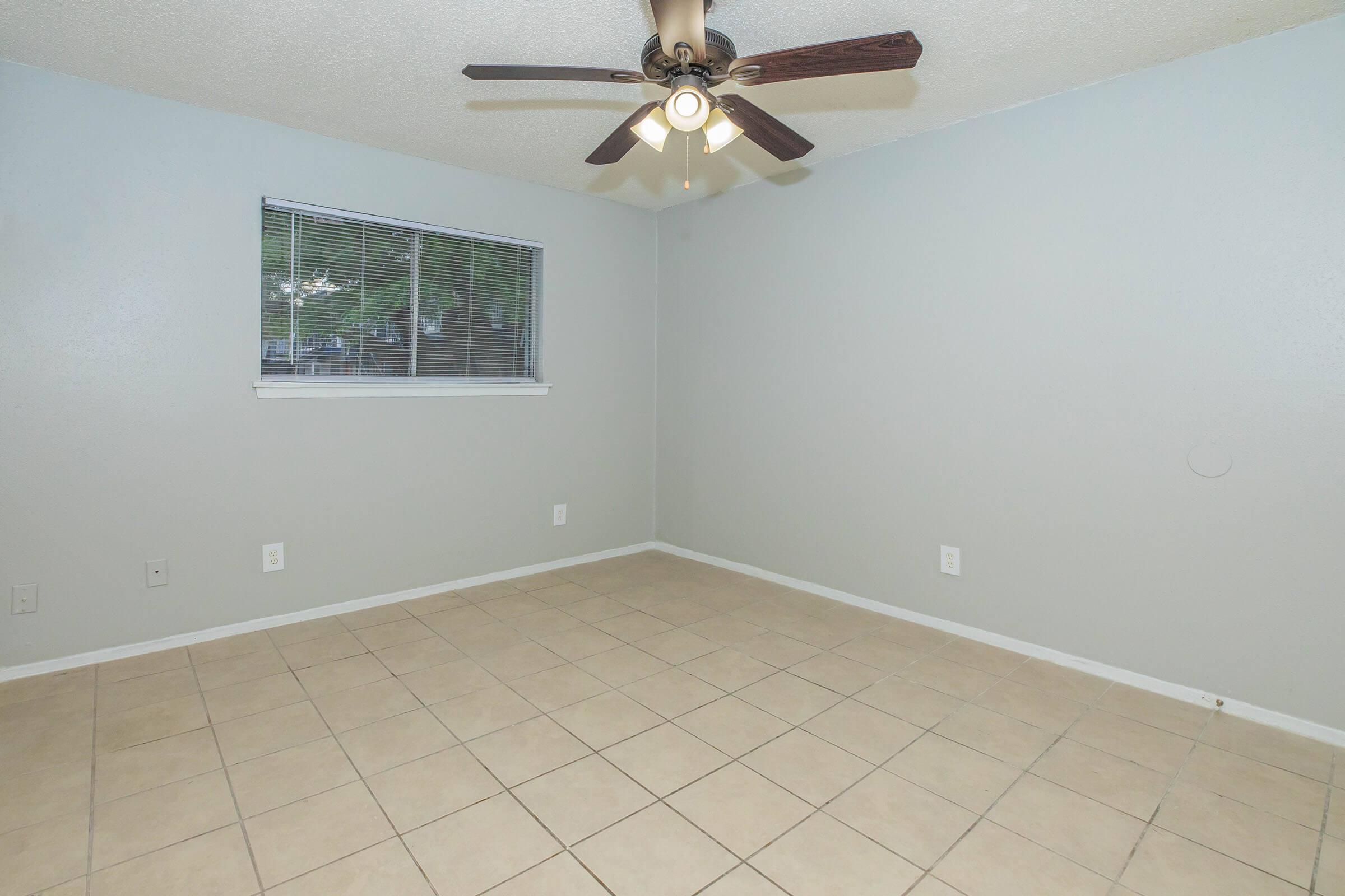 A small, empty room with light gray walls and a tiled floor. There is a window with blinds allowing natural light in, and a ceiling fan with three lights is mounted above. The room has no furniture, creating a spacious and open feel.