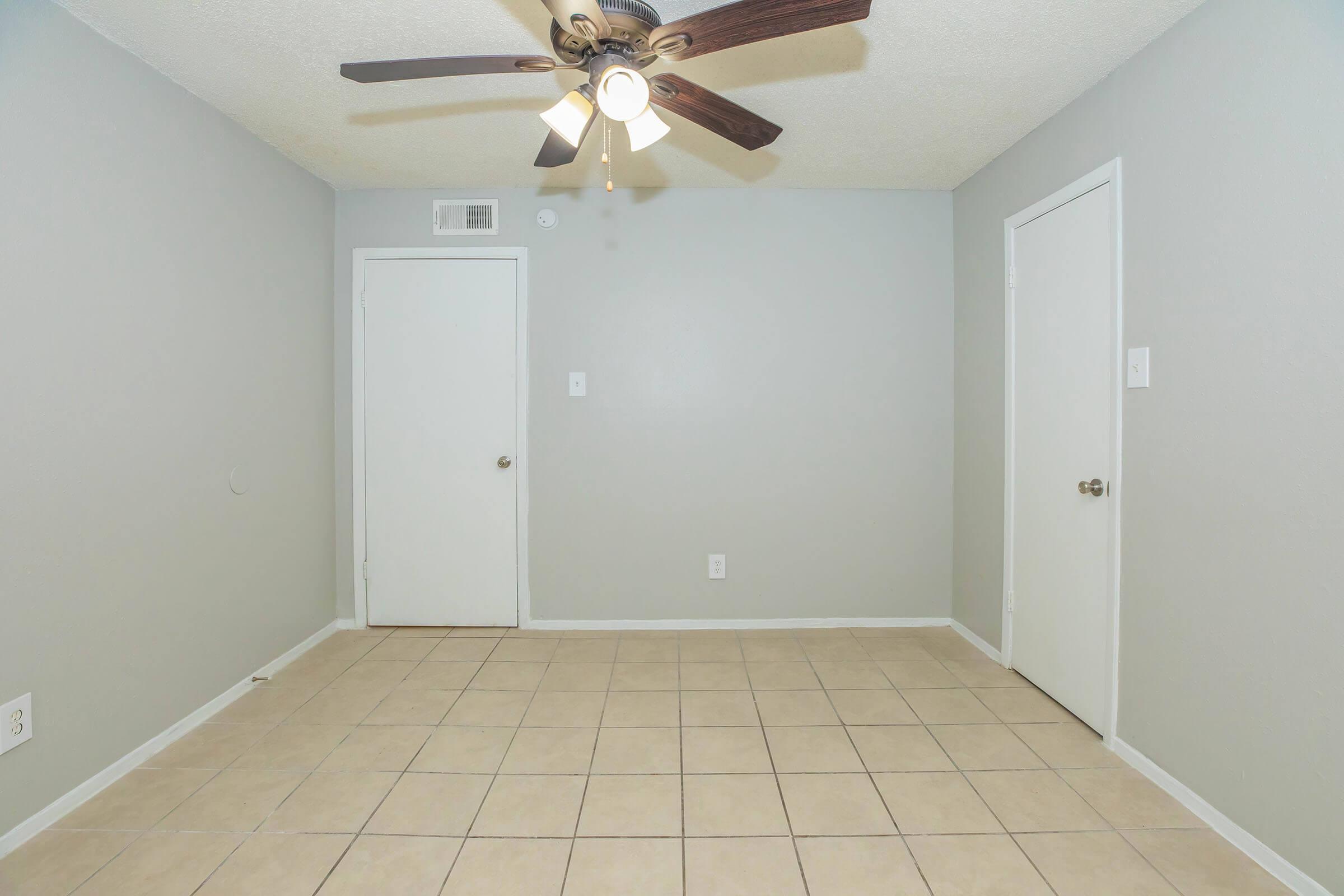 A minimalistic room featuring gray walls, a ceiling fan with wooden blades, and light-colored tile flooring. There are two closed doors, one on the left and one on the right, with no furniture or decorations present, creating a spacious and empty atmosphere.