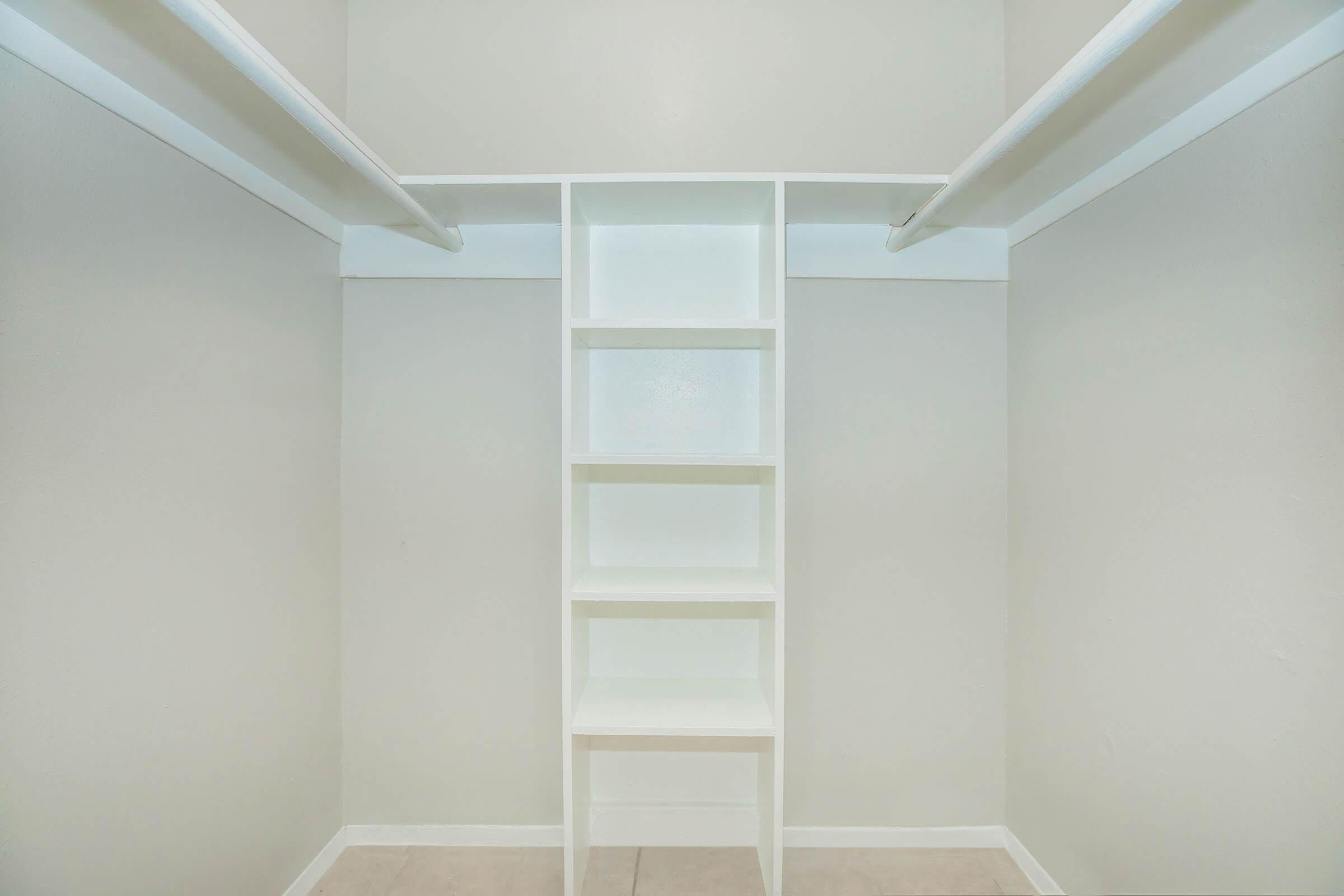 A spacious closet with two hanging rods and a simple white bookshelf in the center. The walls and shelves are painted in a light color, creating a bright and airy atmosphere. The flooring is tiled, enhancing the clean, minimalist look of the space.
