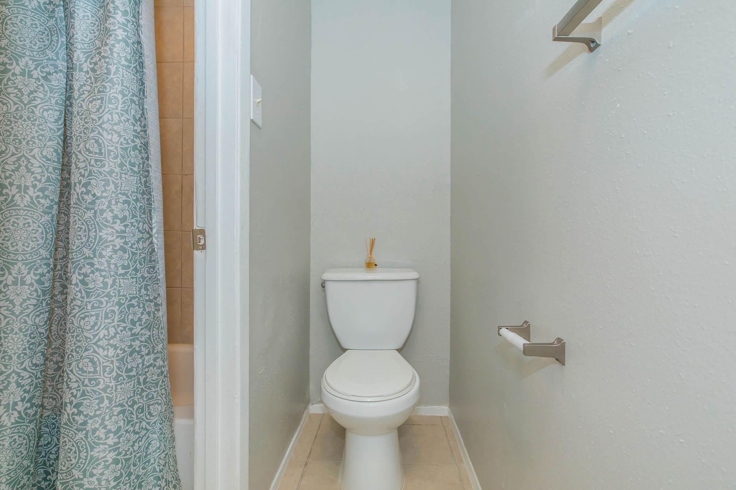 A small bathroom featuring a white toilet, light-colored walls, and a decorative shower curtain. The space is well-lit and includes metal towel racks and a neutral floor tile.