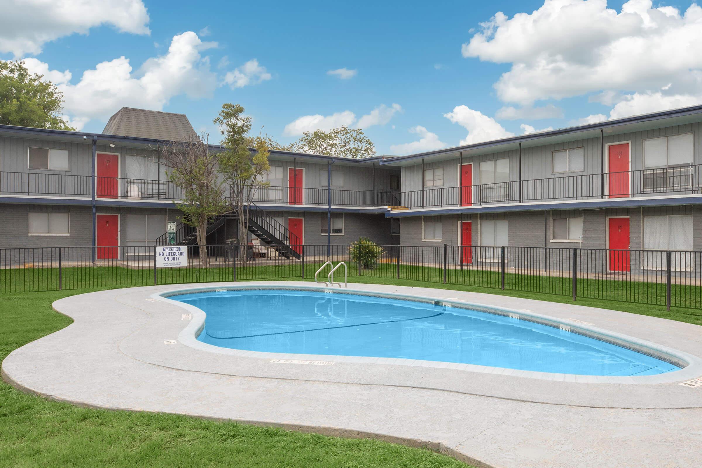 A clear blue swimming pool surrounded by green grass, with a two-story gray apartment building in the background. The building features red doors and a staircase leading to the upper floor. The sky is bright with fluffy white clouds.