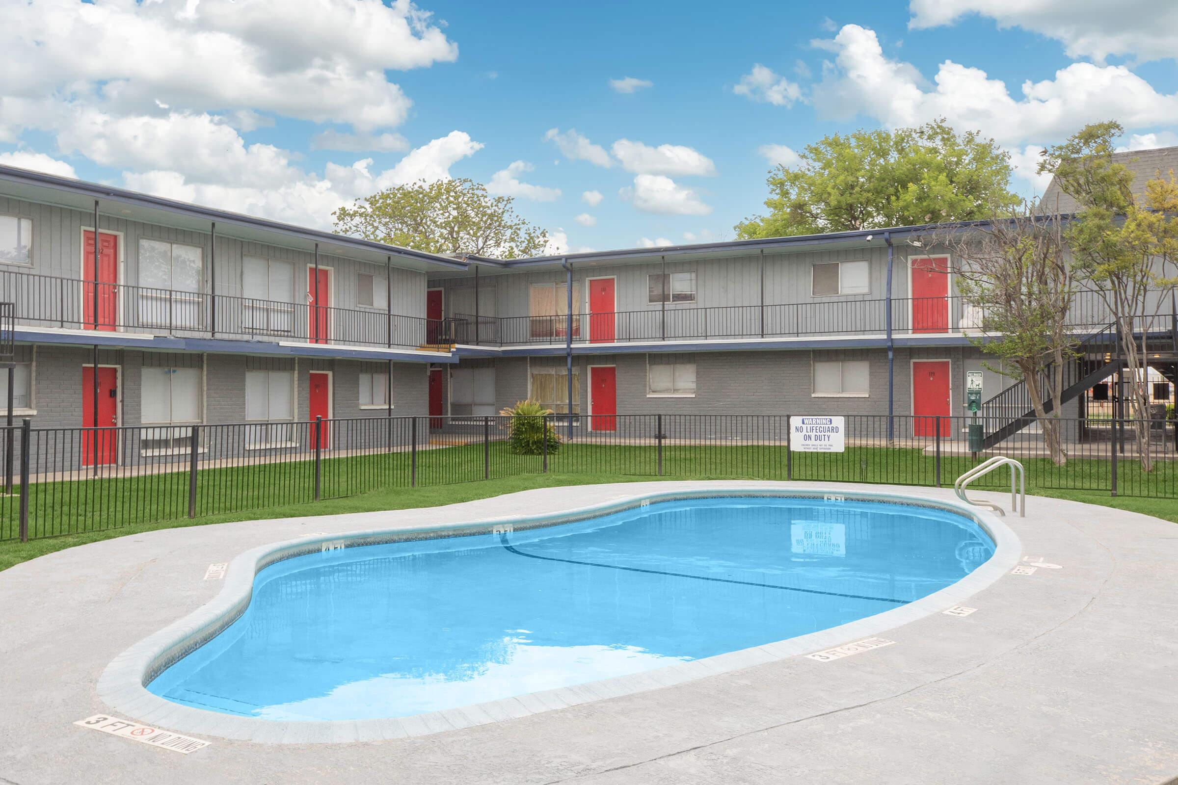 A clear blue pool surrounded by a gated area, with a two-story building in the background featuring gray walls and red doors. Lush green grass and a few trees can be seen, under a partly cloudy sky.