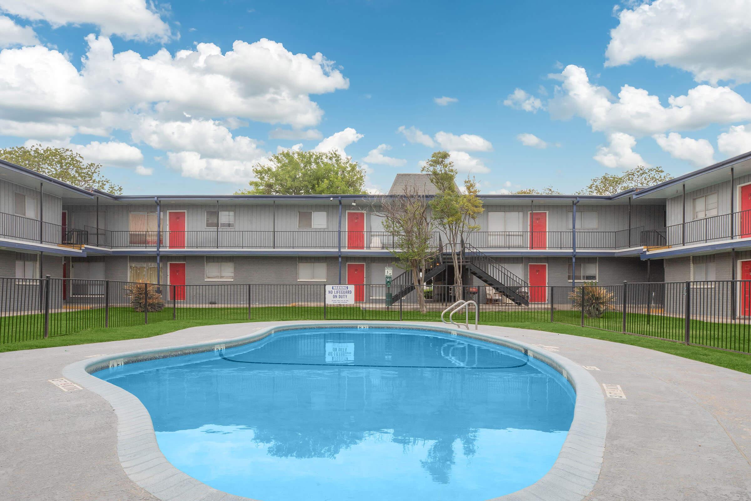 View of a residential courtyard featuring a swimming pool in the foreground. Surrounding the pool are two-story apartment buildings with gray walls and red door accents. Lush green grass is present, along with a few small trees and a clear blue sky filled with fluffy white clouds.