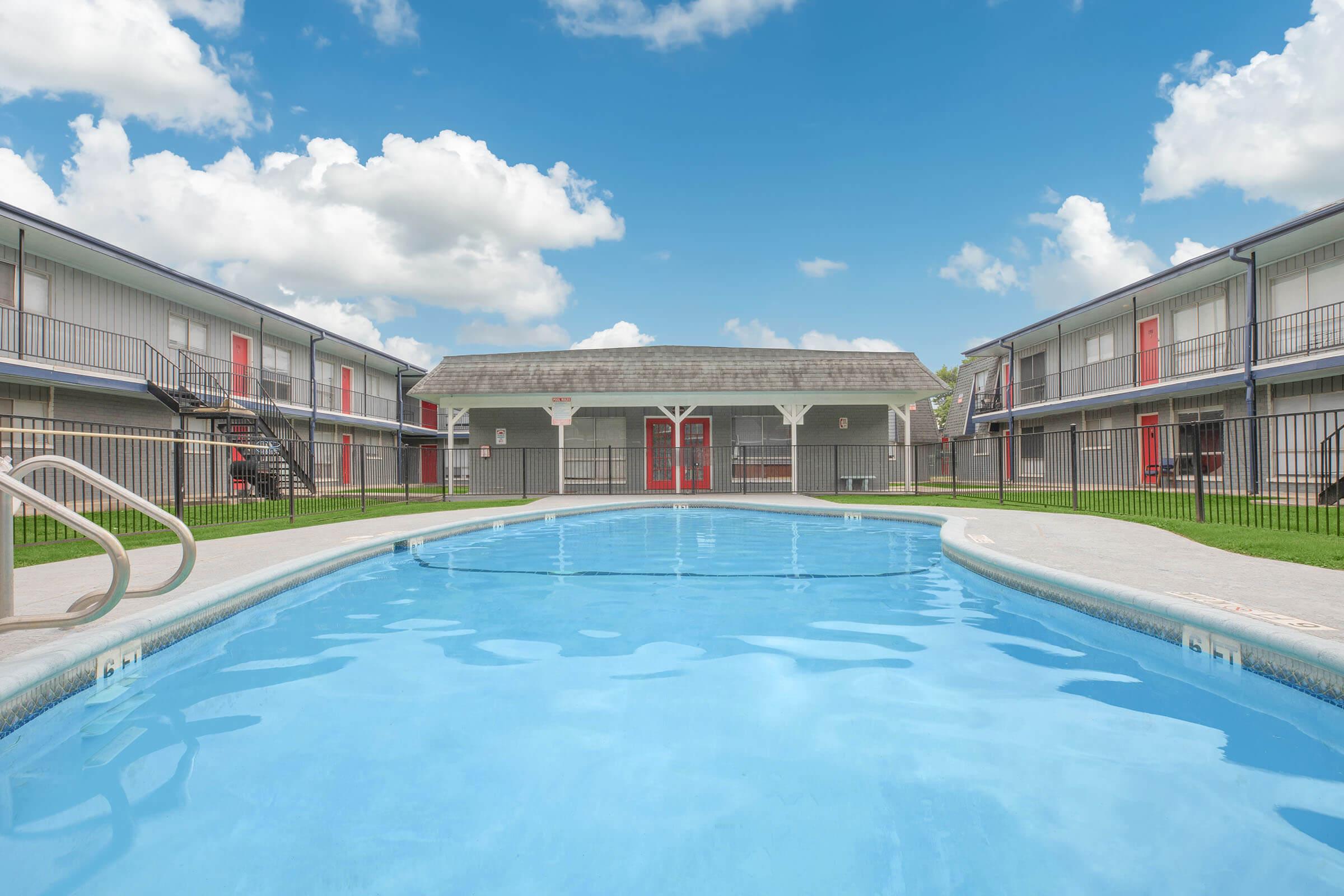 A clear swimming pool in the foreground, surrounded by a lawn and two-story apartment buildings in the background. The buildings are painted in neutral tones with red doors, and there are scattered fluffy clouds in a bright blue sky above.