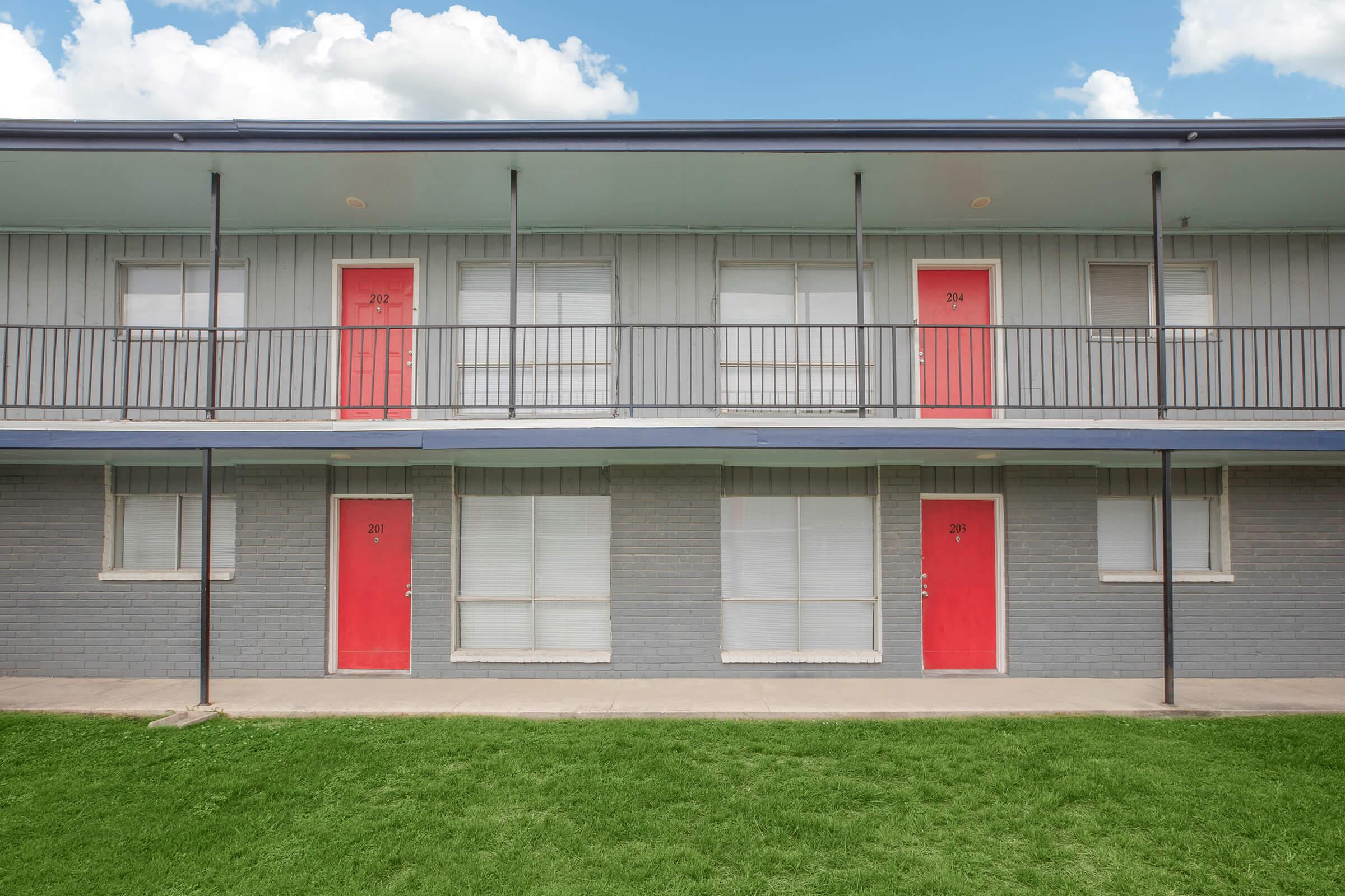 Two-story motel with a gray exterior featuring red doors. The second floor has balconies with black railings, and several windows are covered. A grassy area is in front of the building, under a partly cloudy blue sky.