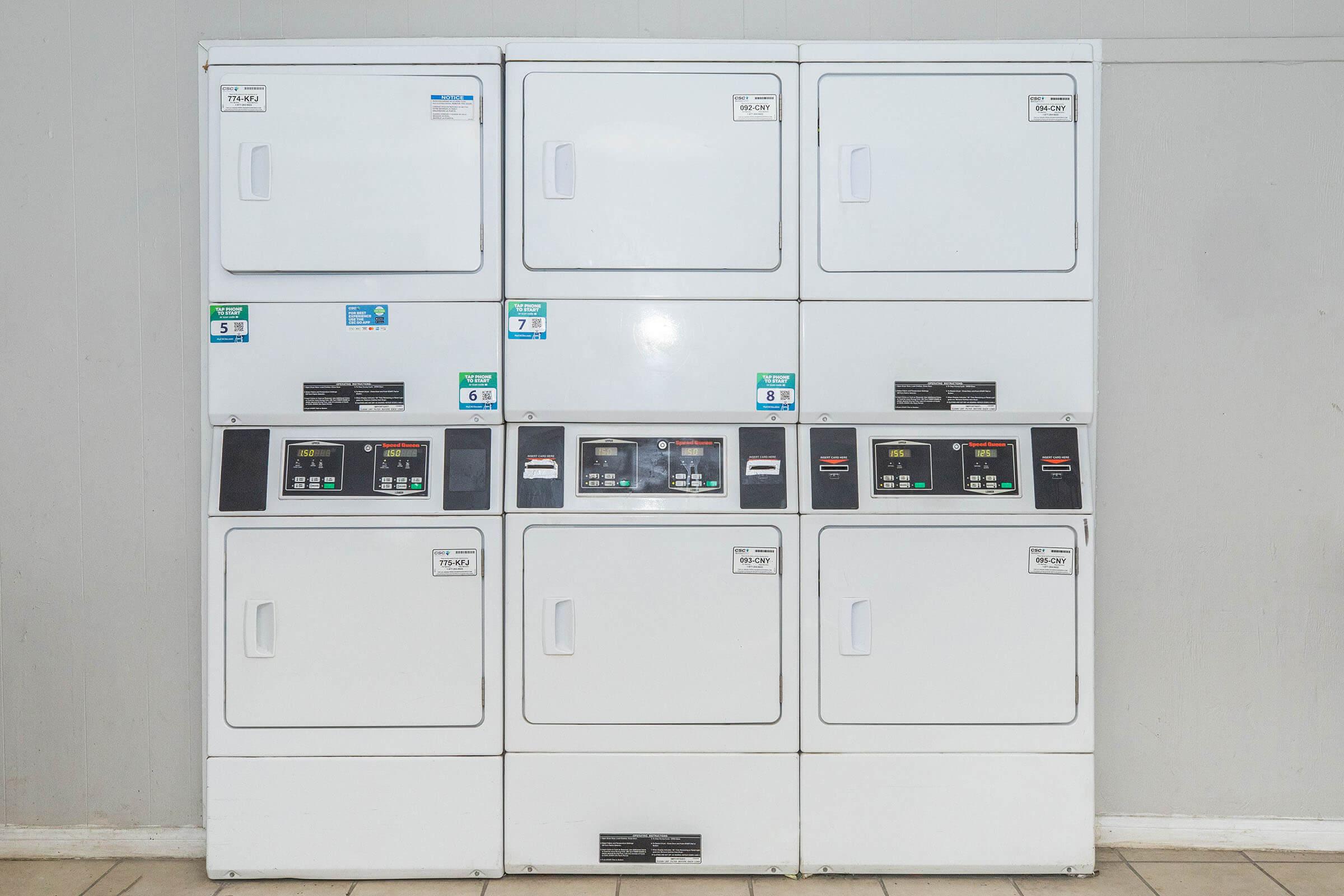 A row of six white commercial washing machines and dryers, stacked in two columns. Each machine features control panels with buttons and displays, along with labels indicating different settings. The wall behind them is light-colored and there is a tiled floor beneath the machines.