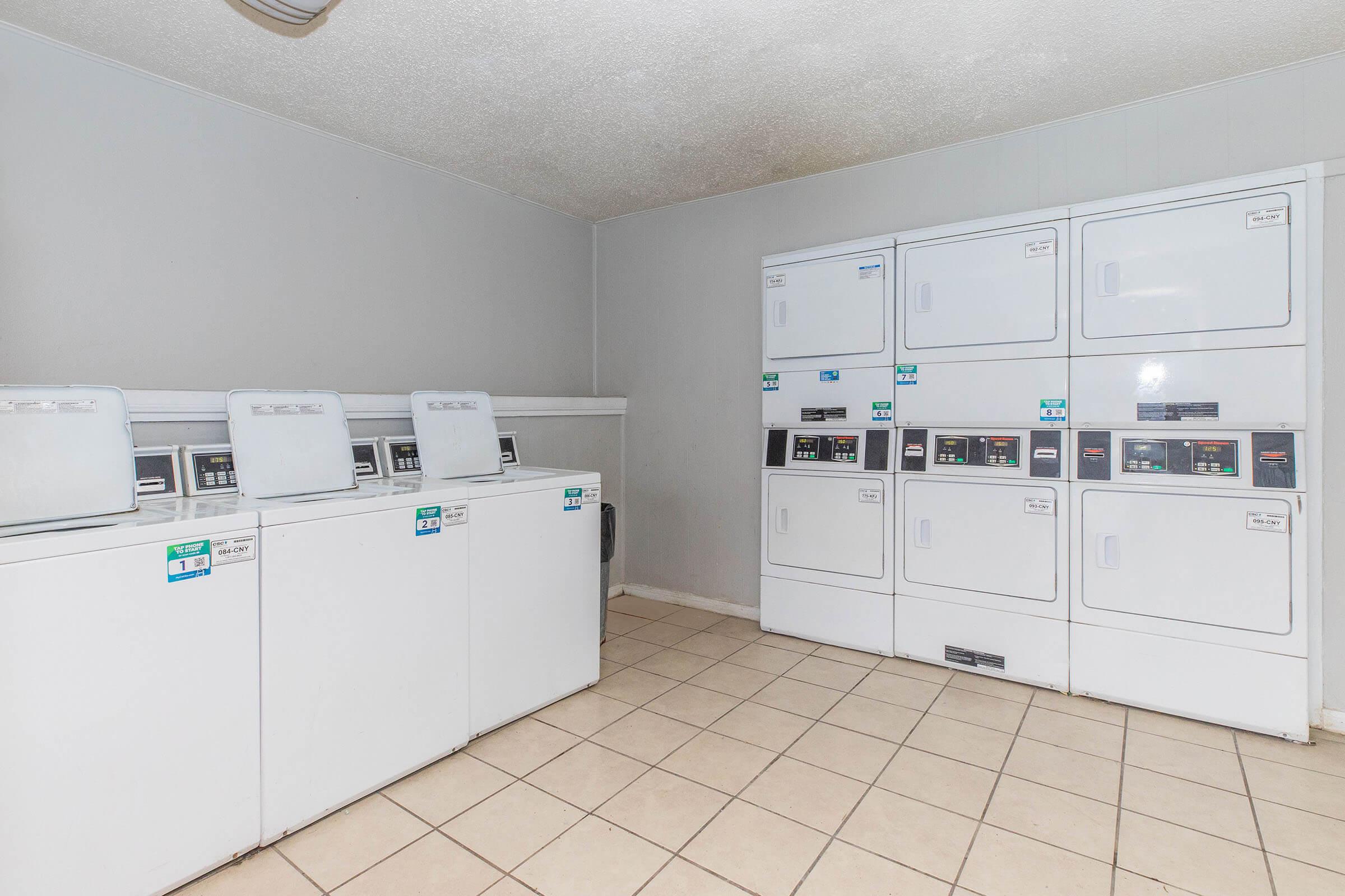 A clean and well-lit laundry room featuring several white washing machines and dryers arranged neatly against the wall. The floor is tiled, and the walls are painted a light color, creating a tidy and organized space for laundry activities.