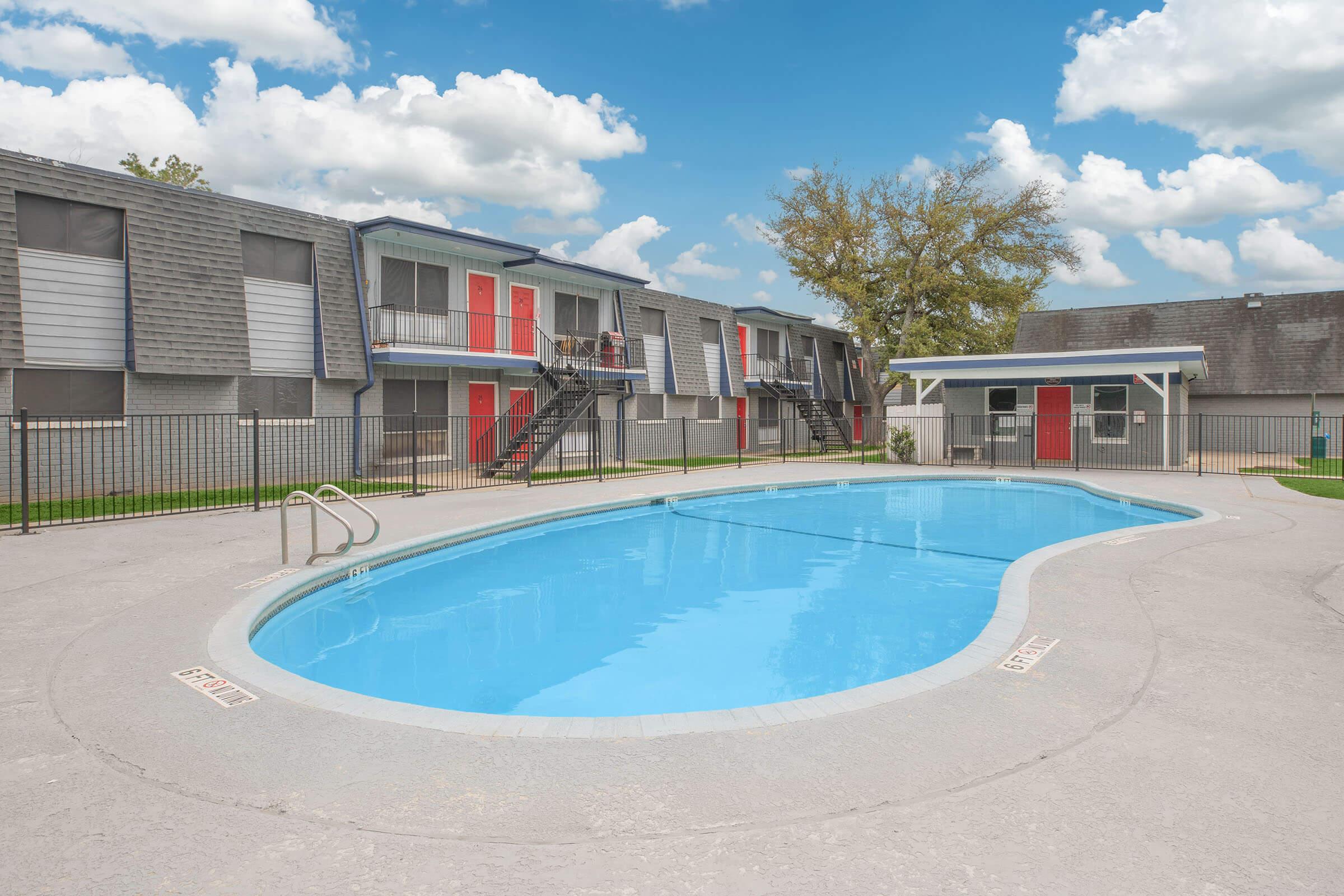 A bright outdoor swimming pool surrounded by gray apartment buildings with red accents. The pool area is fenced, featuring a gently sloped entry. A pavilion with a red door is visible nearby, and the sky is partly cloudy, adding a serene atmosphere to the scene.