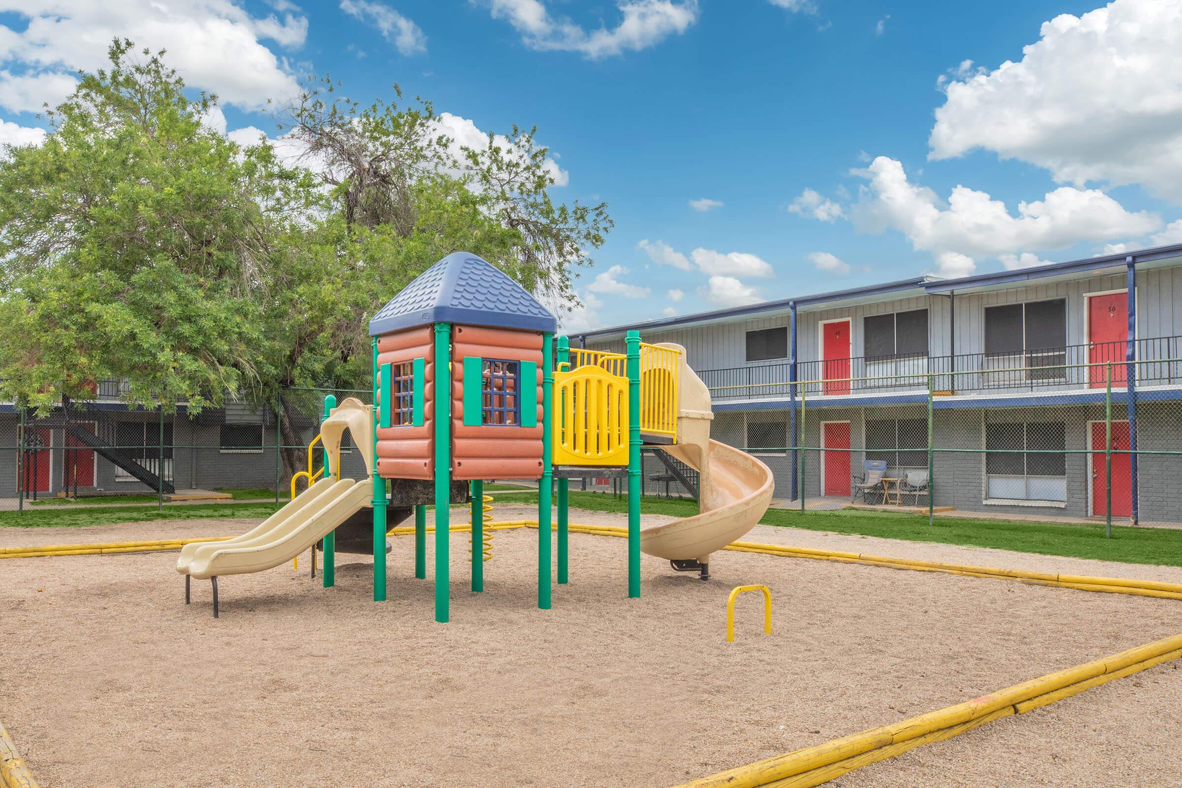 A colorful playground structure with slides and climbing features, situated in a sandy area. In the background, there are several apartments with red doors, framed by trees and a blue sky with fluffy clouds.