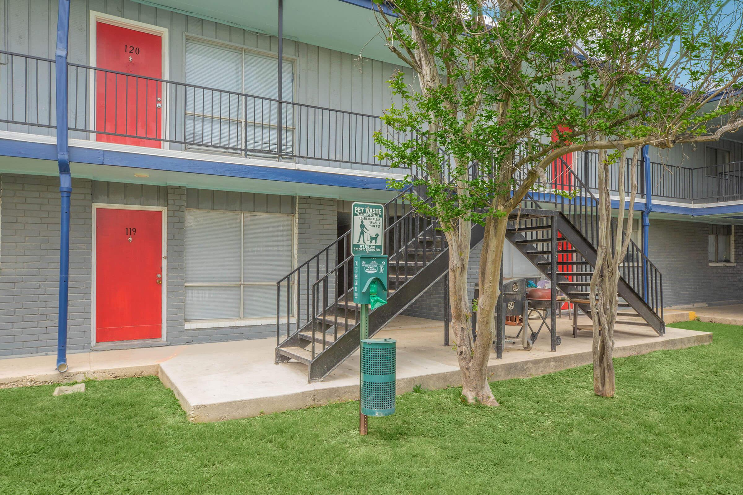 Two-story apartment building with red doors labeled 119 and 120. A set of black stairs leads to the second floor. In front, there's a green dog waste station near a grassy area. Trees provide some shade, and the building exterior features a gray brick design with blue accents.