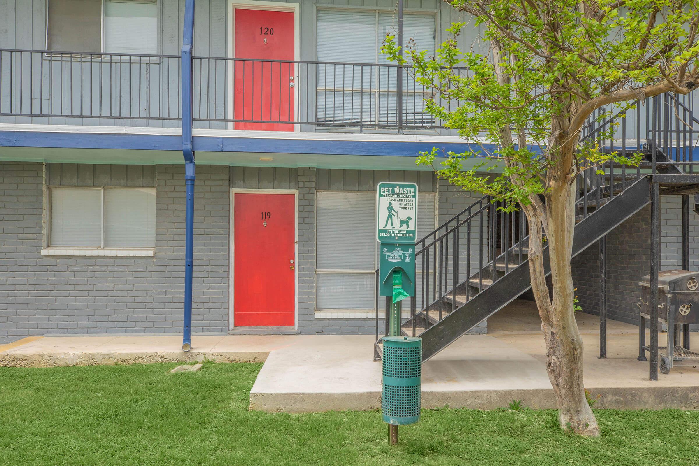 A two-story building with blue siding featuring two red doors numbered 120 and 119. A green pet waste station is placed beside a tree in the front yard, with a staircase leading to the second floor. The area is well-maintained with grass and concrete pathways.