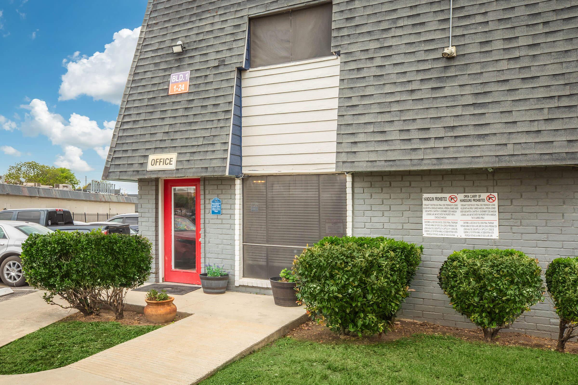 A gray building with a sloped roof and a red door labeled "OFFICE." The entrance features a screen door and is flanked by two potted plants. In front, there is a walkway leading to the door. To the right, a sign displays building regulations. The background includes a blue sky with clouds.