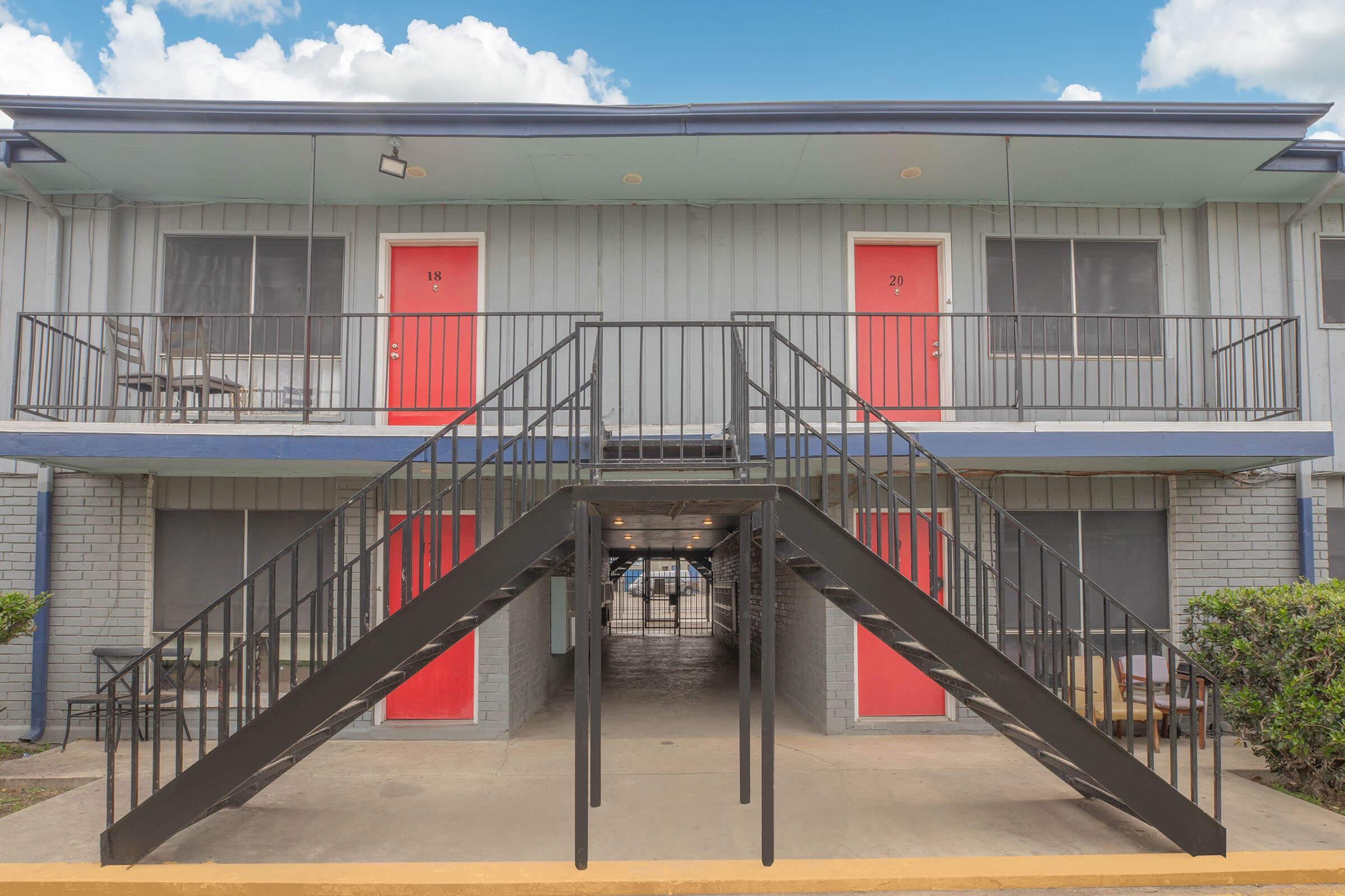 Exterior view of a two-story apartment building featuring multiple units. The building has a gray exterior with two prominent red doors numbered 18 and 20. A black metal staircase leads to the upper level, and a covered pathway connects the units. The sky above is partly cloudy.