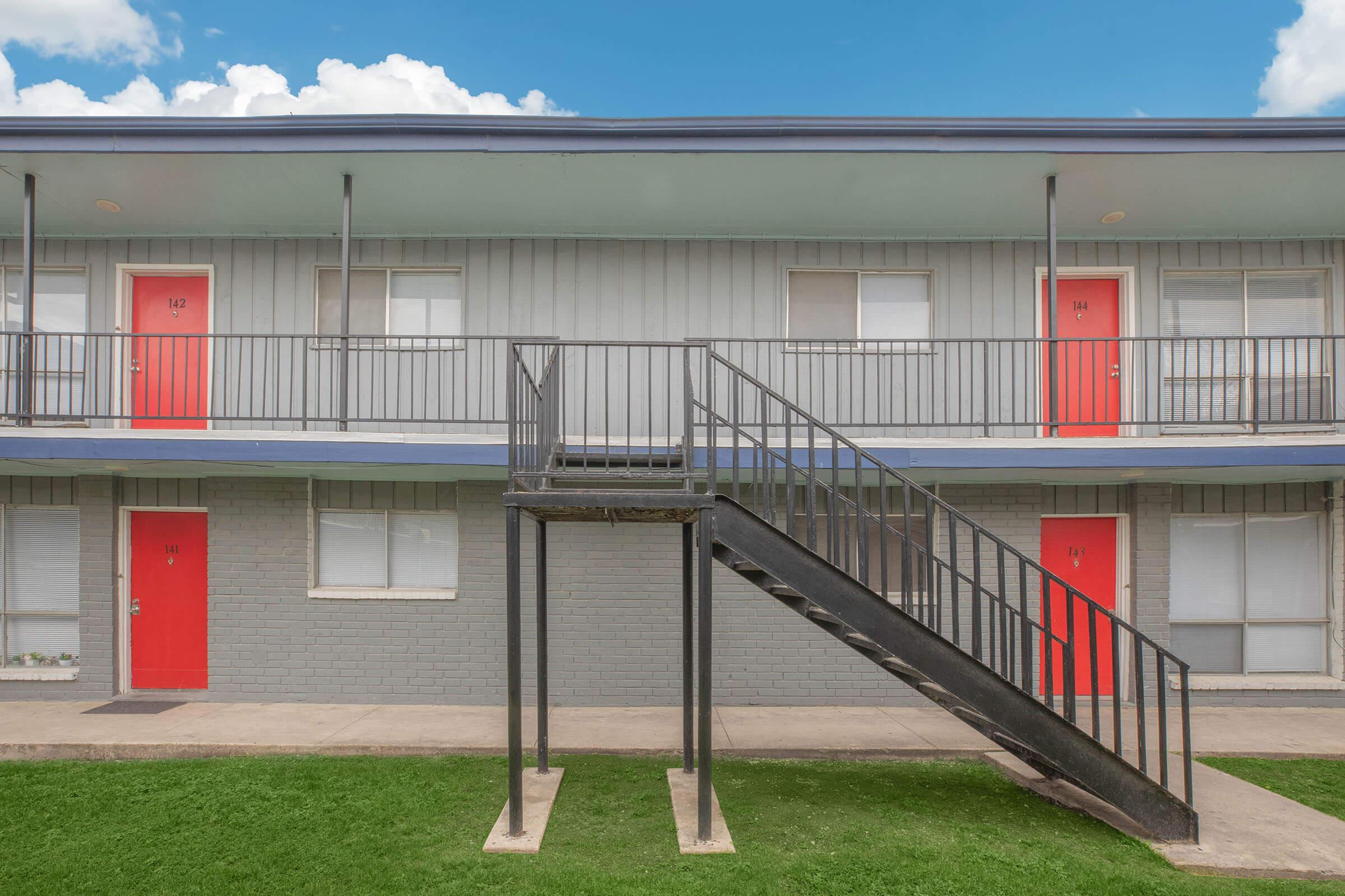 A view of a two-story apartment building with gray siding and bright red doors. An outdoor staircase leads to the upper level. Green grass is visible in the foreground, and the sky is partly cloudy.