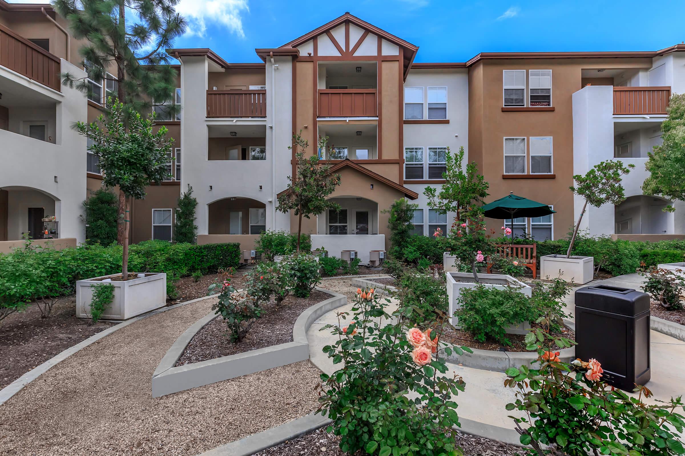 A landscaped courtyard with pathways bordered by plants and flowers, leading to a multi-story apartment building. The building features balconies and large windows, with shaded seating areas visible. Lush greenery surrounds the space, creating a peaceful outdoor environment.