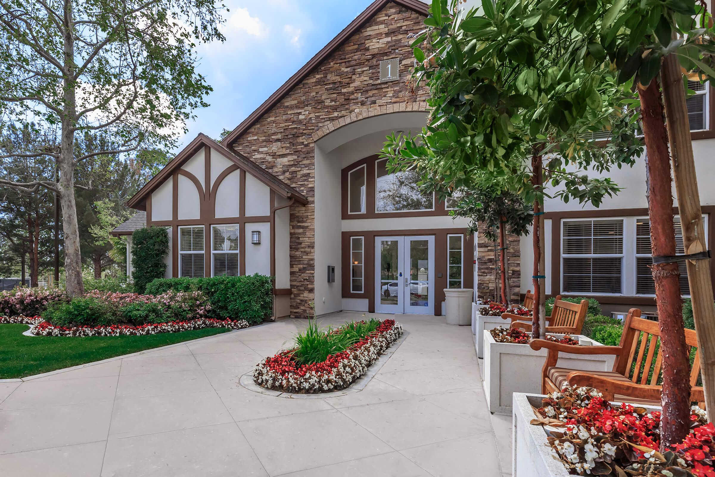 A landscaped entrance to a building featuring stone accents and large windows. Flanking the entrance are well-maintained flowerbeds with colorful blooms, and there are wooden benches on either side, providing a welcoming outdoor space. Trees and greenery enhance the inviting atmosphere.