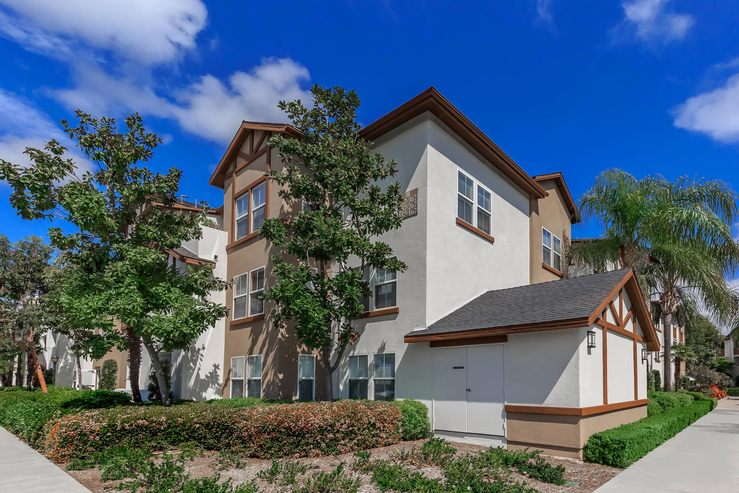 A modern two-story apartment building with a light-colored exterior and brown accents, surrounded by well-maintained landscaping. The sky is bright blue with scattered clouds, and there are palm trees in the vicinity. The setting appears sunny and inviting, ideal for residential living.