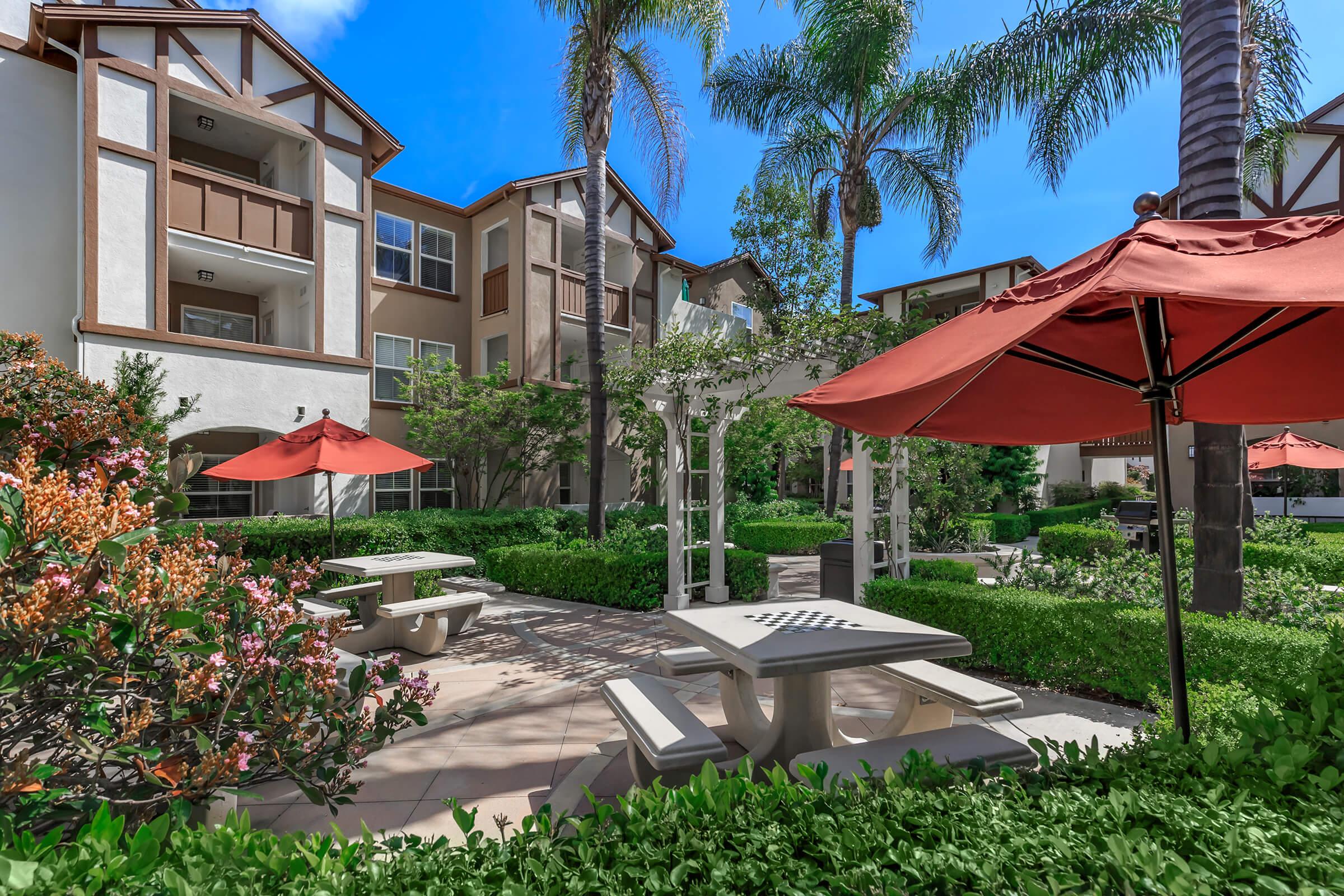 A landscaped courtyard featuring picnic tables, red umbrellas, and well-maintained greenery. Surrounding the area are residential buildings with balconies, and palm trees casting shadows in the sunlight. The sky is blue with a few clouds, creating a bright, inviting outdoor space.