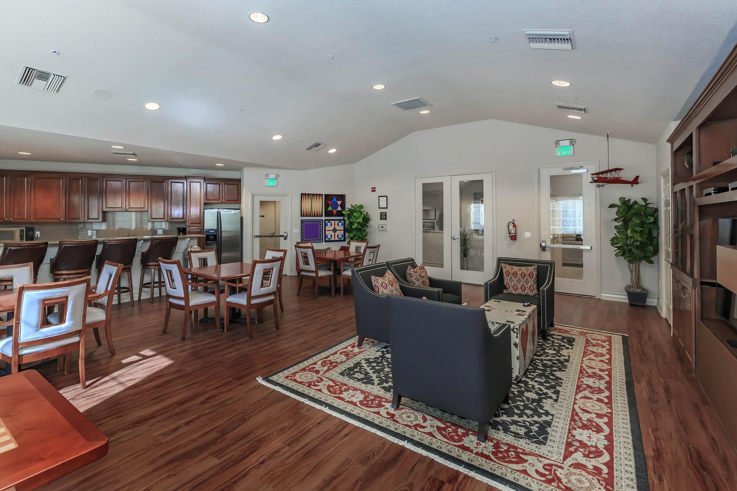 A spacious common area featuring dining tables and chairs, a cozy seating arrangement with sofas, hardwood flooring, and a large rug. The room has large windows and modern décor, including a kitchenette area with wooden cabinets and a few decorative items on the walls.