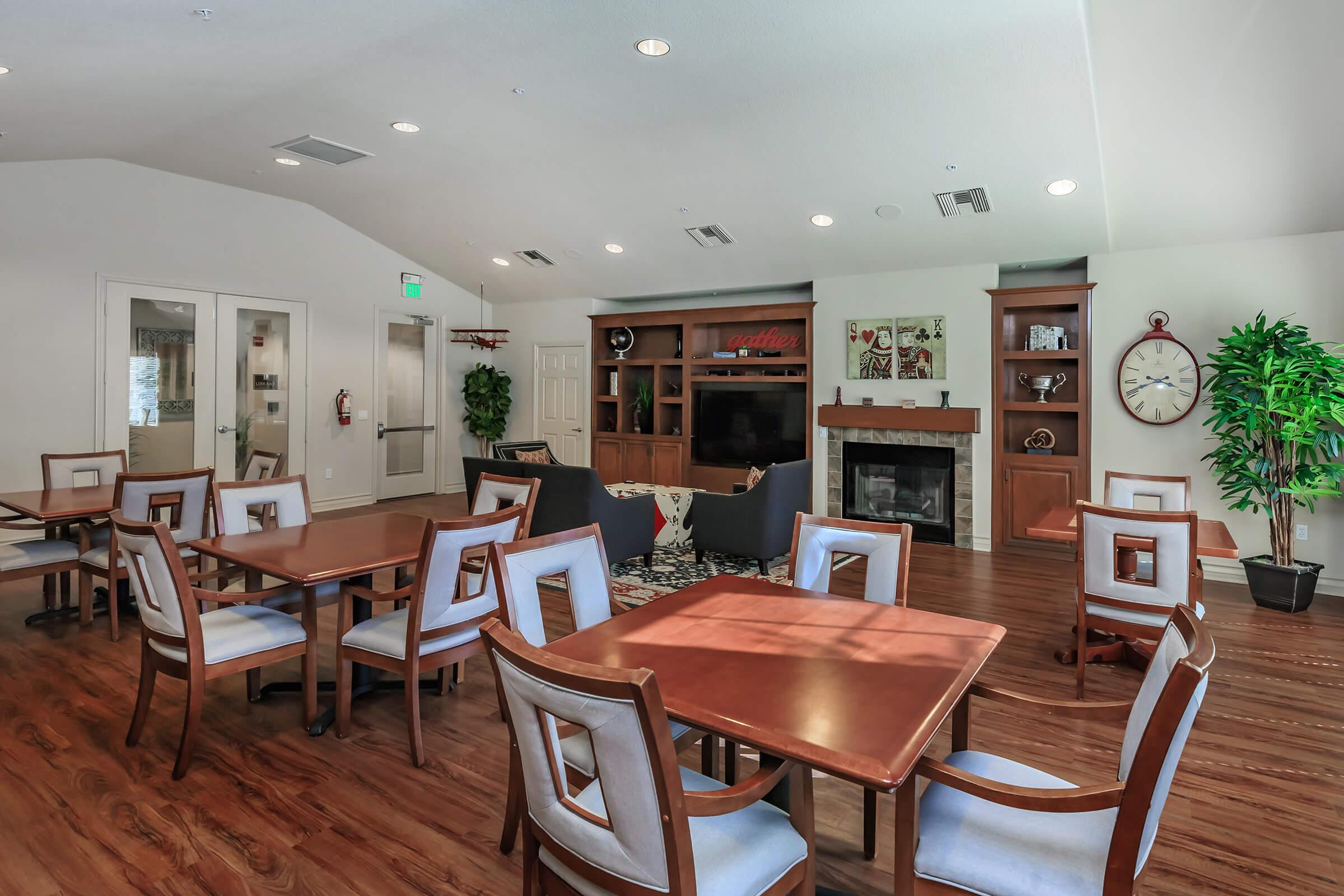 A spacious common area featuring wooden dining tables and chairs, a cozy seating arrangement with sofas, a warm fireplace, and decorative shelves. Natural light fills the room, enhancing the inviting atmosphere, perfect for social gatherings or relaxation.