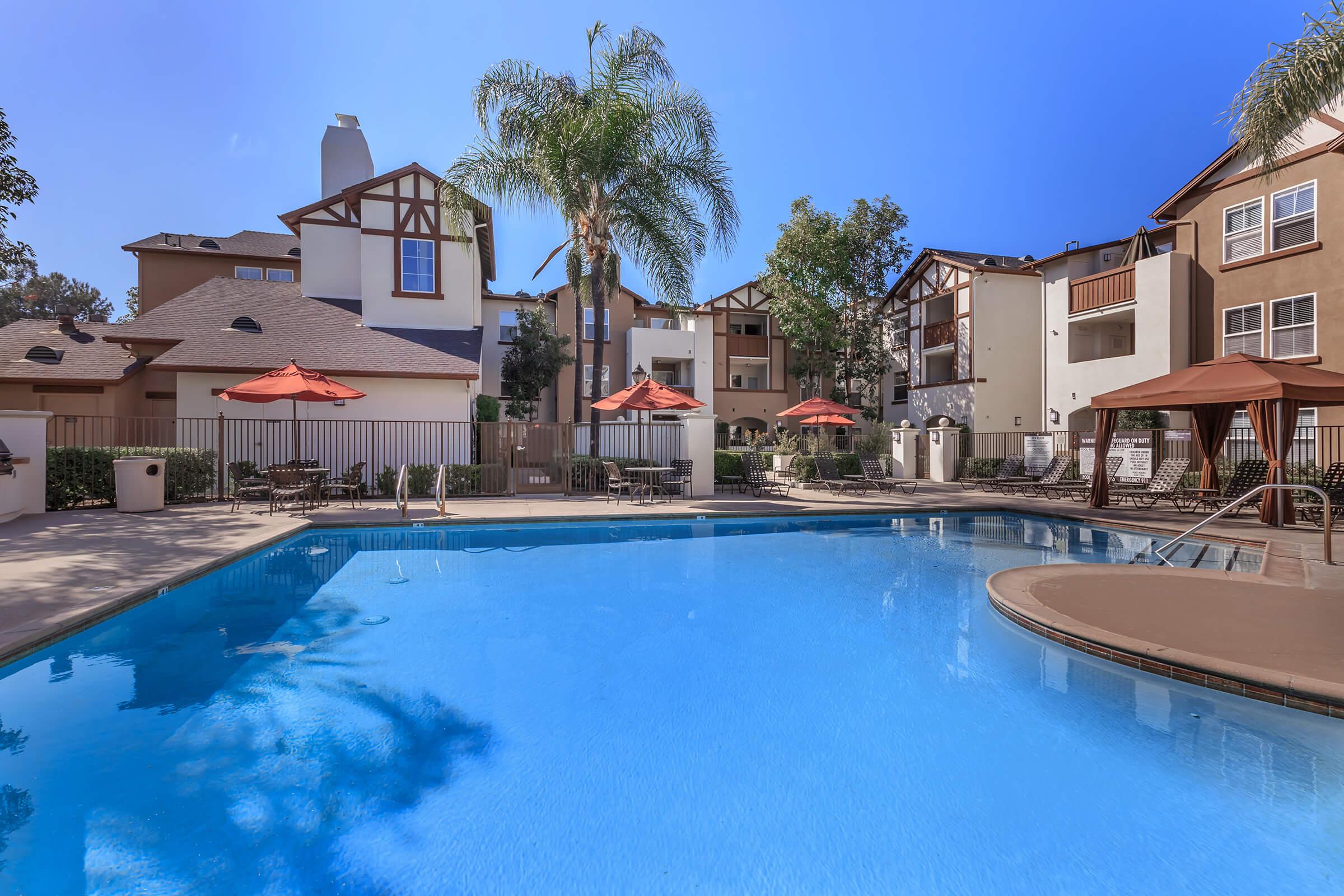 A clear blue swimming pool surrounded by lounge areas and umbrellas, with palm trees and apartment buildings in the background. The scene is sunny and inviting, featuring a well-maintained outdoor space ideal for relaxation.