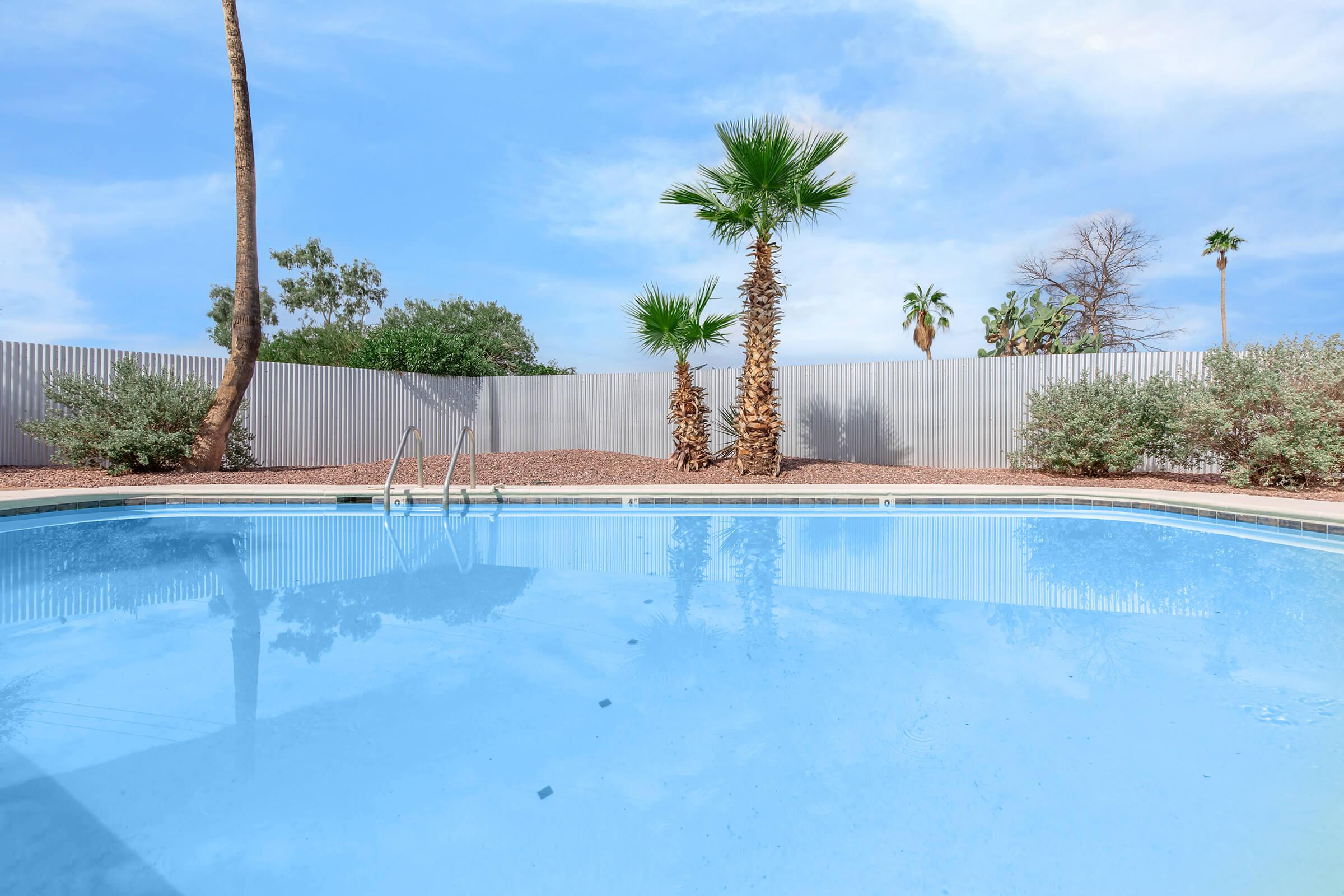 A clear blue swimming pool surrounded by a light-colored fence. Palm trees and other greenery are visible in the background under a bright blue sky with scattered clouds. The pool area is clean and inviting, suitable for relaxation and leisure activities.