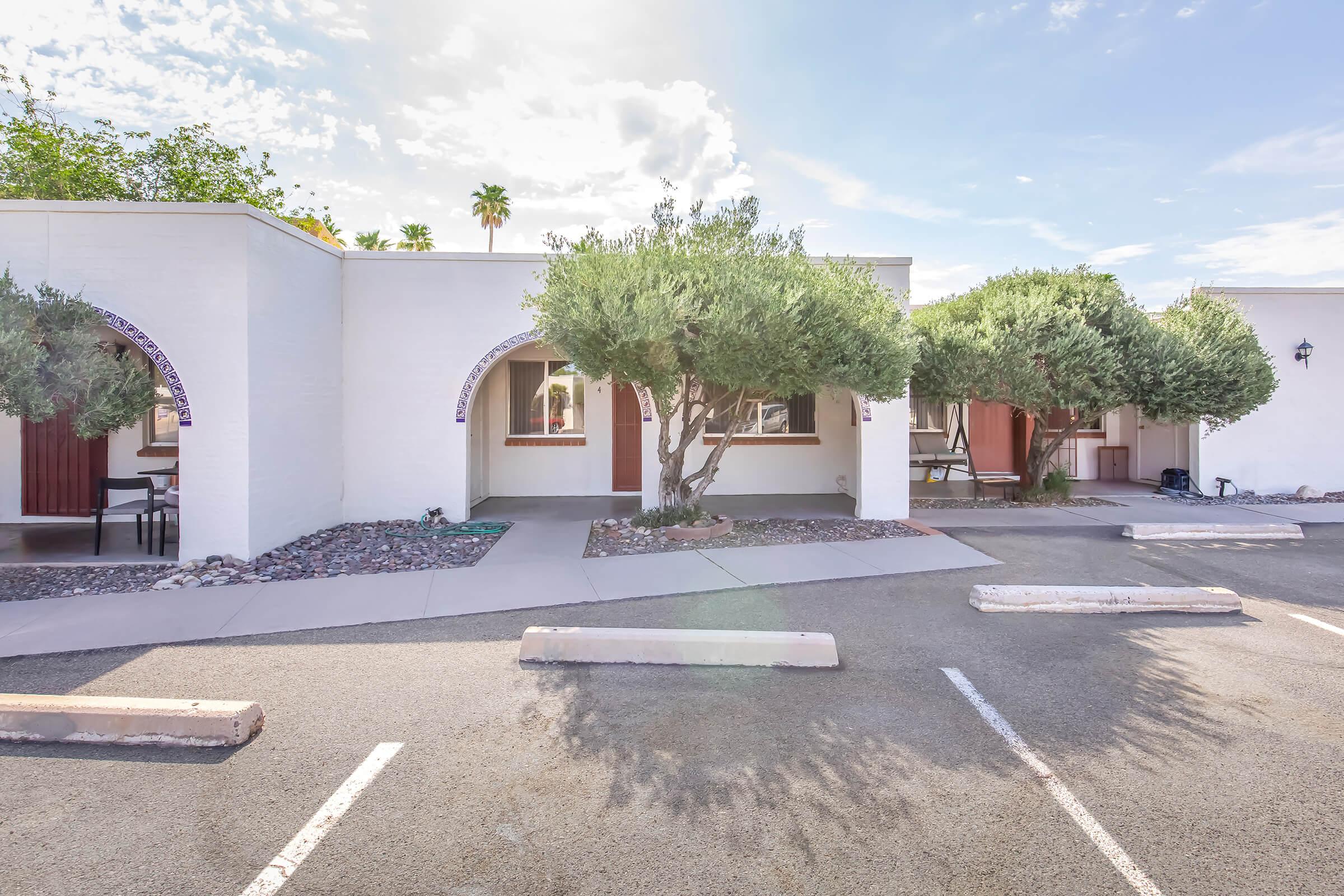 A view of a light-colored building with arched doorways surrounded by greenery. Two olive trees are planted in front, and there are parking spaces visible in the foreground. The sky is partly cloudy with a hint of sunlight.