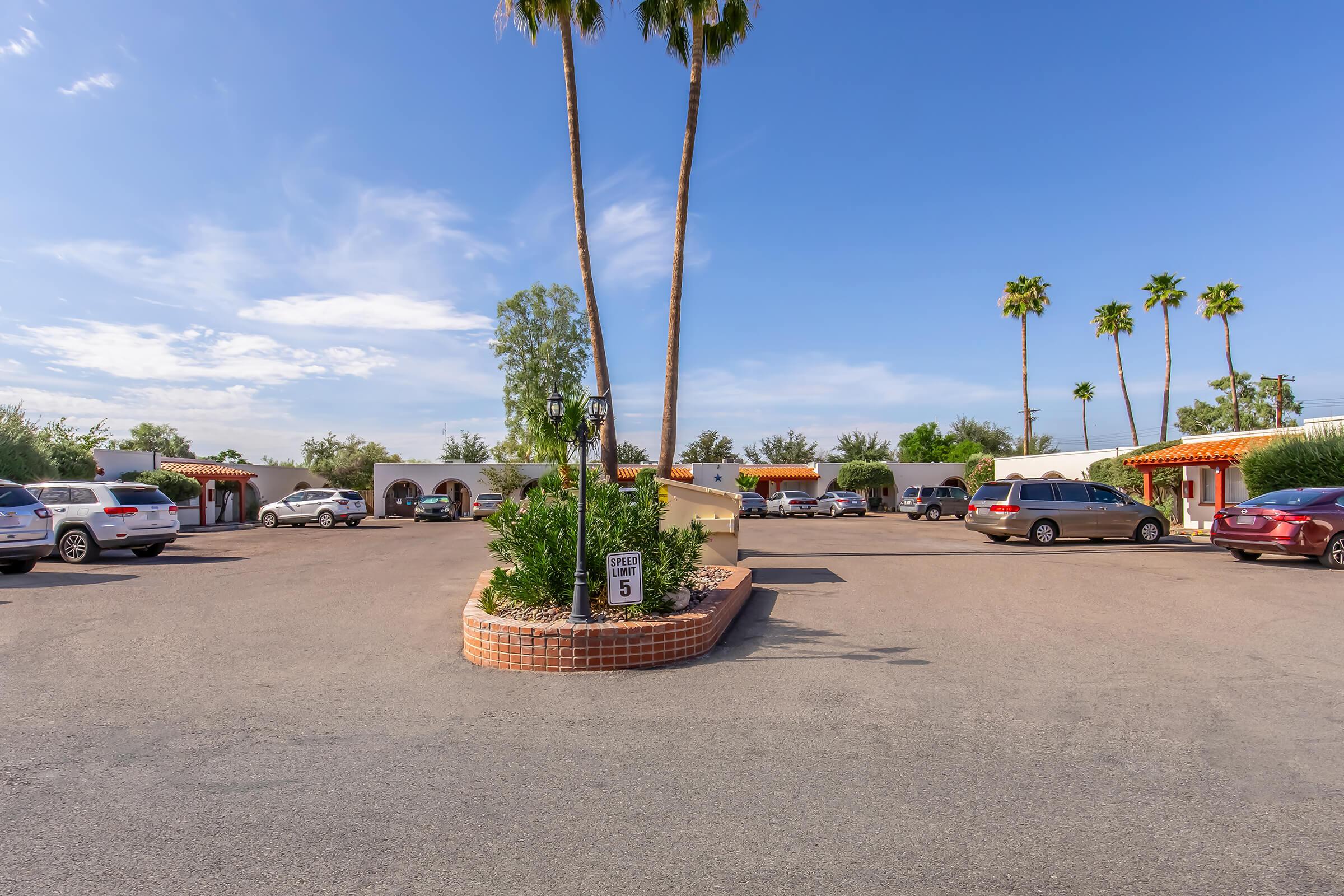 A parking lot featuring several parked cars, palm trees, and a landscaped area with circular planting. The sky is mostly clear with a few clouds, and the surrounding buildings have a Southwestern architectural style. A parking space sign is visible in the foreground.