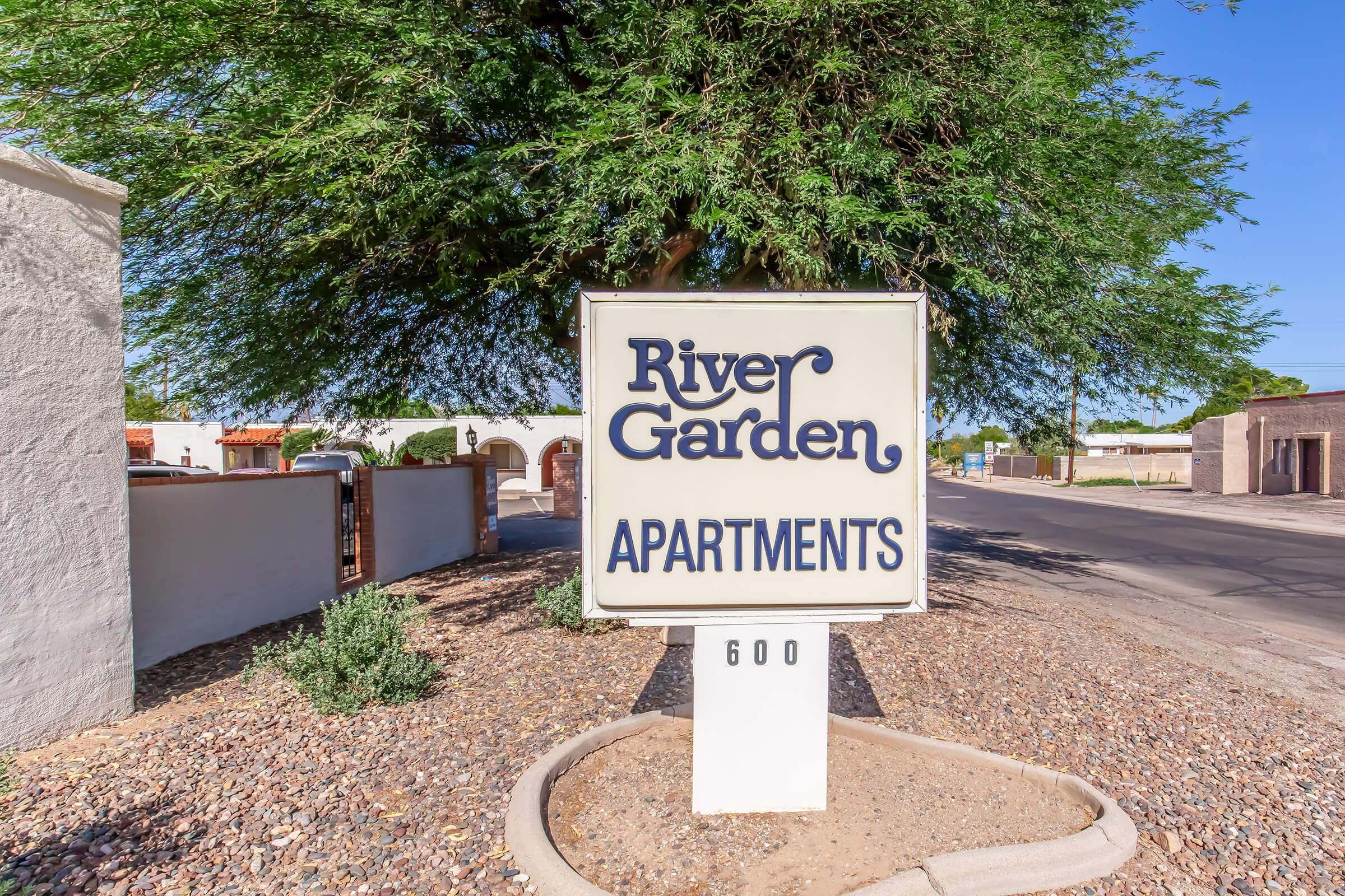 Sign for River Garden Apartments, featuring the name in an elegant font. The sign is white with blue text, situated near a landscaped area with a tree and gravel. The address "600" is displayed at the bottom, indicating the location of the apartments along a roadside.
