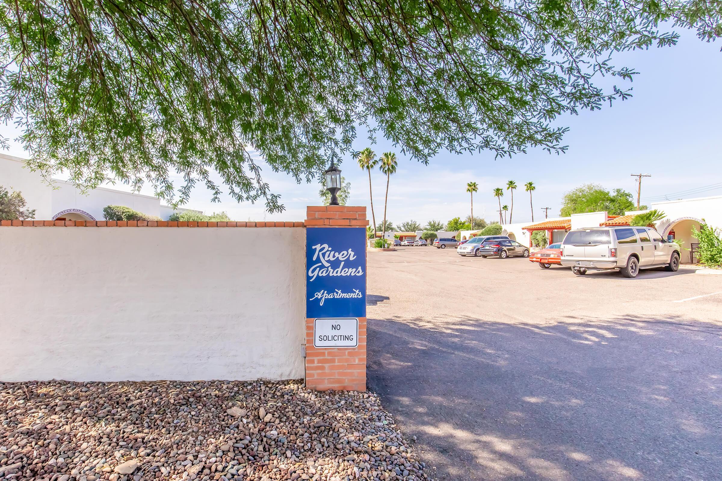 Sign for River Gardens Apartments, with a "No Soliciting" notice, next to a parking lot. The area is bordered by a low wall and surrounded by palm trees, under a clear blue sky.
