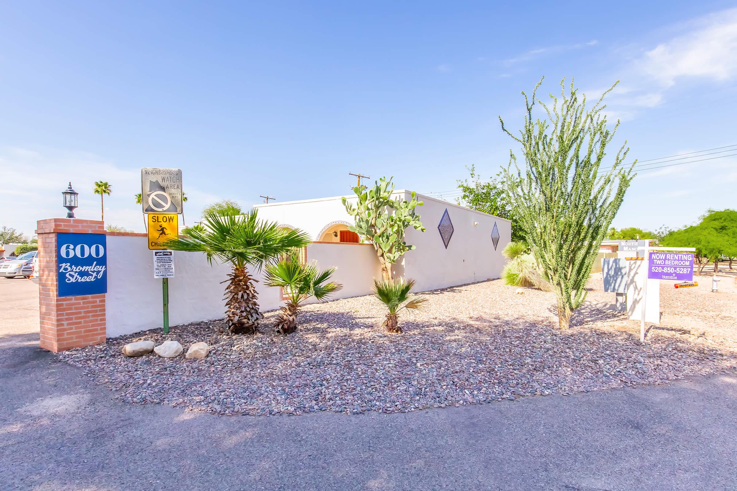 A residential building with a white stucco exterior, surrounded by desert landscaping including palm trees and cacti. There is a brick sign displaying the address "600 Bansley Street." A traffic sign indicates "SLOW" and there is a real estate sign in front of the property. The sky is clear and sunny.