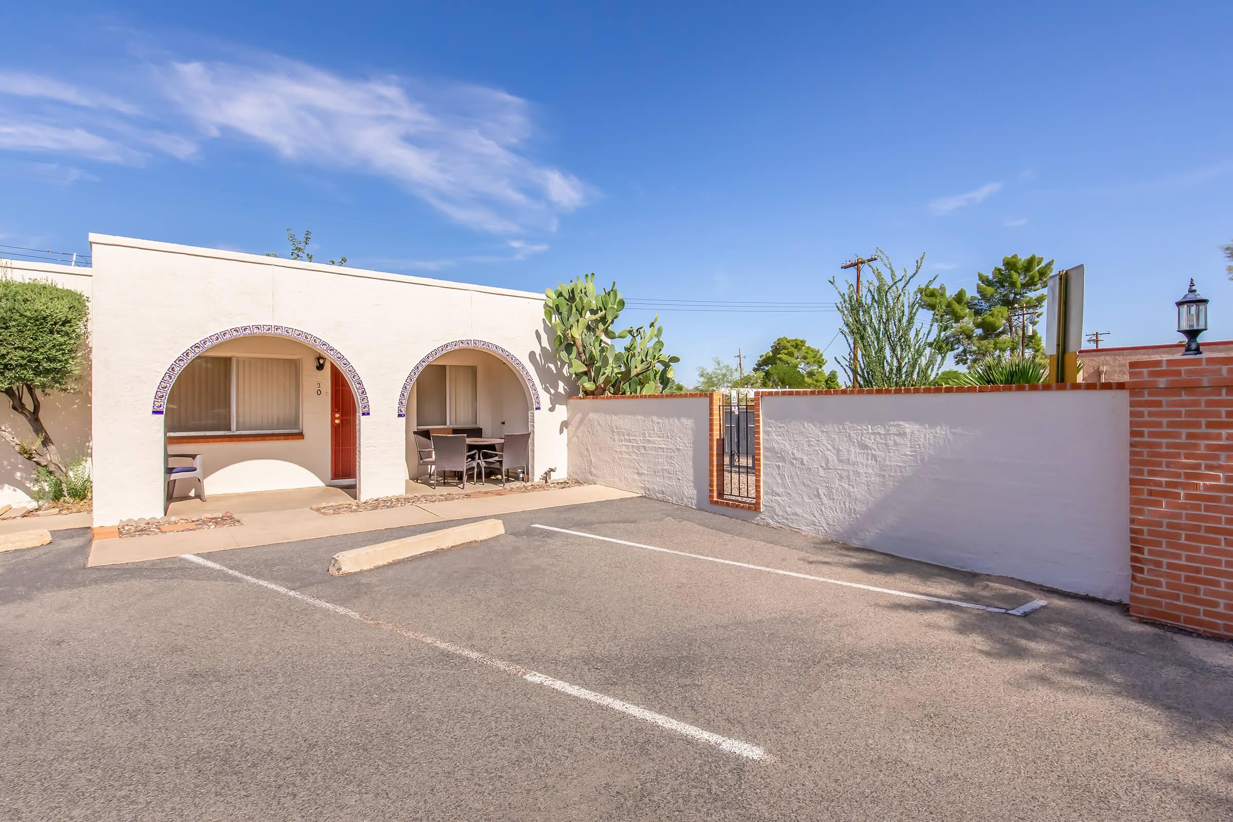 A light-colored building with arched doorways is shown, along with a small outdoor seating area. There's a parking lot with two empty spaces and a gated entrance. The scene is set against a clear blue sky, with some greenery and desert plants visible nearby.