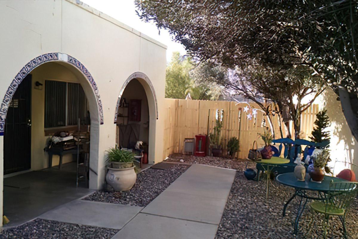Description of a small outdoor area with a pathway leading to a home. There are two arched entryways and a fenced yard featuring trees and colorful decorations. A table and chairs are set up outside, and a few potted plants are visible, creating a charming and inviting atmosphere.