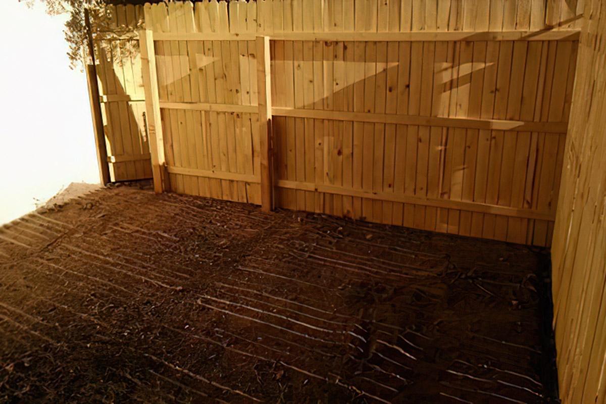 A wooden backyard fence casting shadows on the ground. The area is clear of vegetation, revealing dirt with faint lines or marks. The sunlight creates a pattern of light and shadow across the fence and the ground, showcasing the textures of the wood and earth.