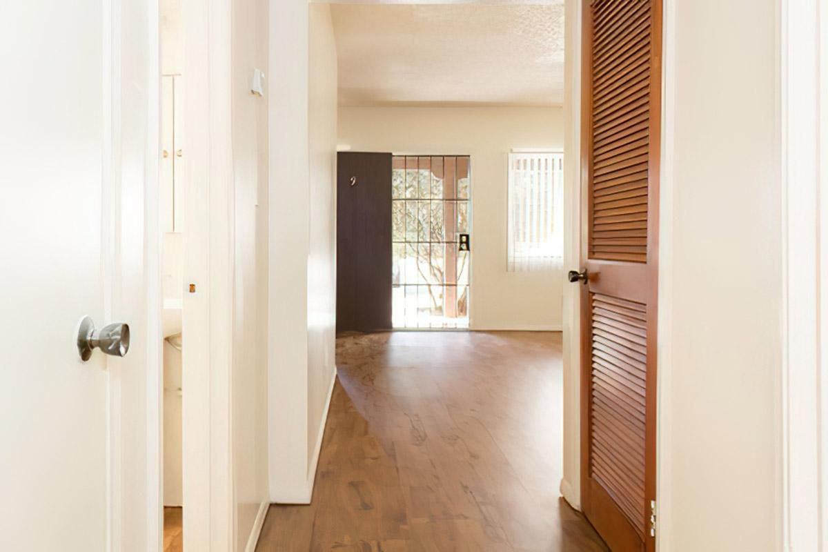 View of a light-colored hallway leading to a living area with wooden flooring. A door with horizontal slats is visible on the right, while the doorway at the end opens to an outdoor area with a screen door and a window letting in natural light. The walls are painted in neutral tones.